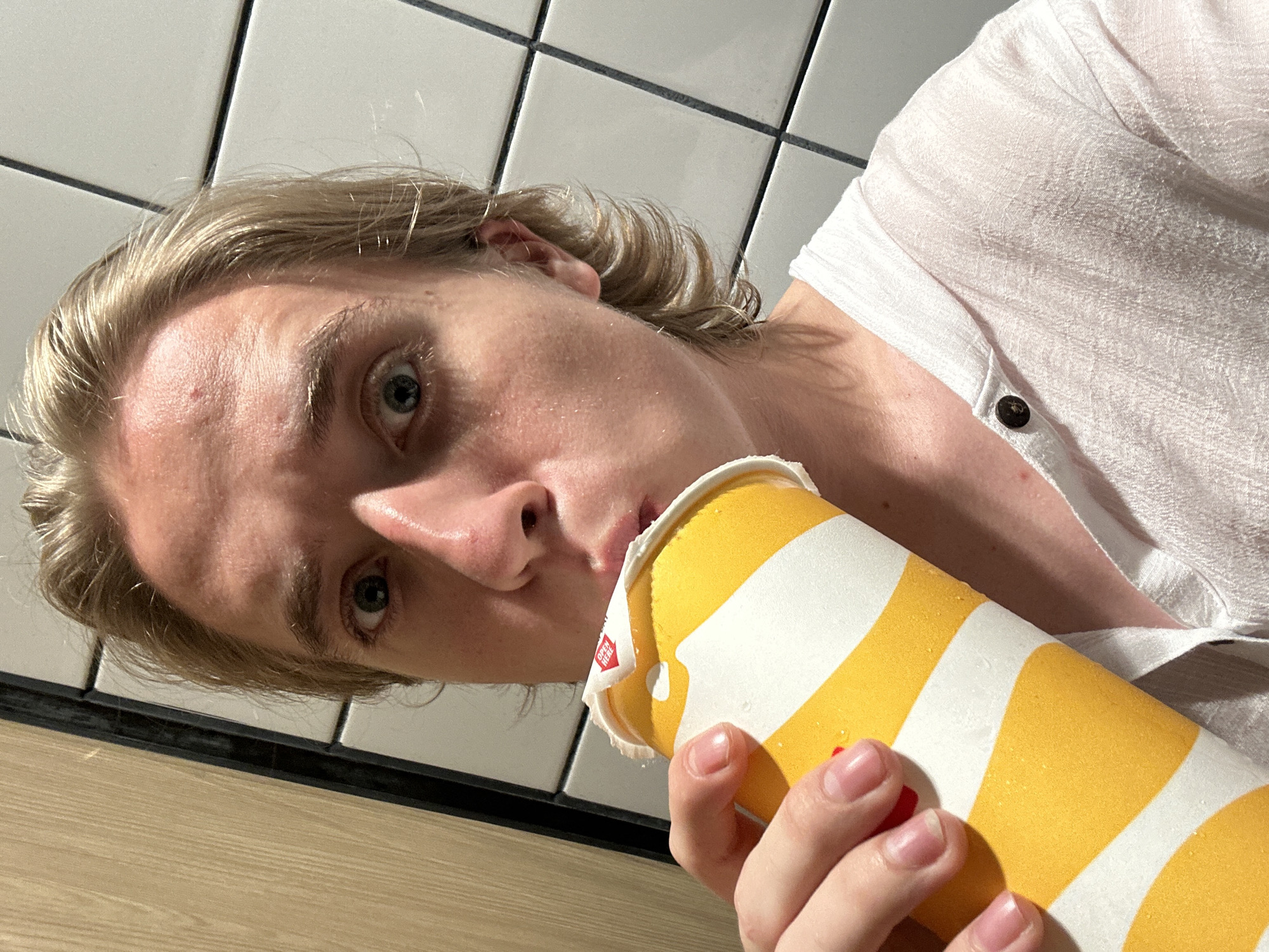 A person with blonde hair and a light-colored shirt is drinking from a yellow and white striped cup. The person's face is partially obscured by the cup. The background consists of tiled walls and a wooden panel. The scene appears to be indoors, possibly in a public restroom or a similar establishment in Thủ Đức, Vietnam.