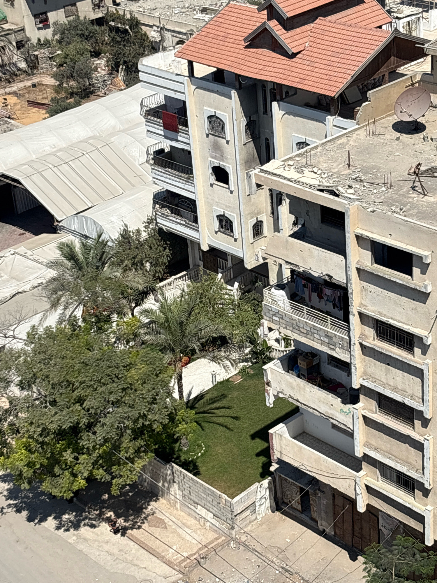 An aerial view captures damaged buildings in the city of Gaza, Palestinian Territories. The scene depicts multi-story residential buildings, one with a red-tiled roof, and another with a damaged concrete rooftop bearing a satellite dish. Several balconies show signs of wear and tear, with laundry hanging on lines on one. Below, a patch of green lawn is visible, bordered by a low brick wall. A large, leafy tree casts a shadow on a paved area where a person in striped clothing is seated. The surrounding urban landscape shows other structures, some appearing to be under construction or in a state of disrepair. The overall impression is one of a densely populated area with signs of ongoing development and damage.