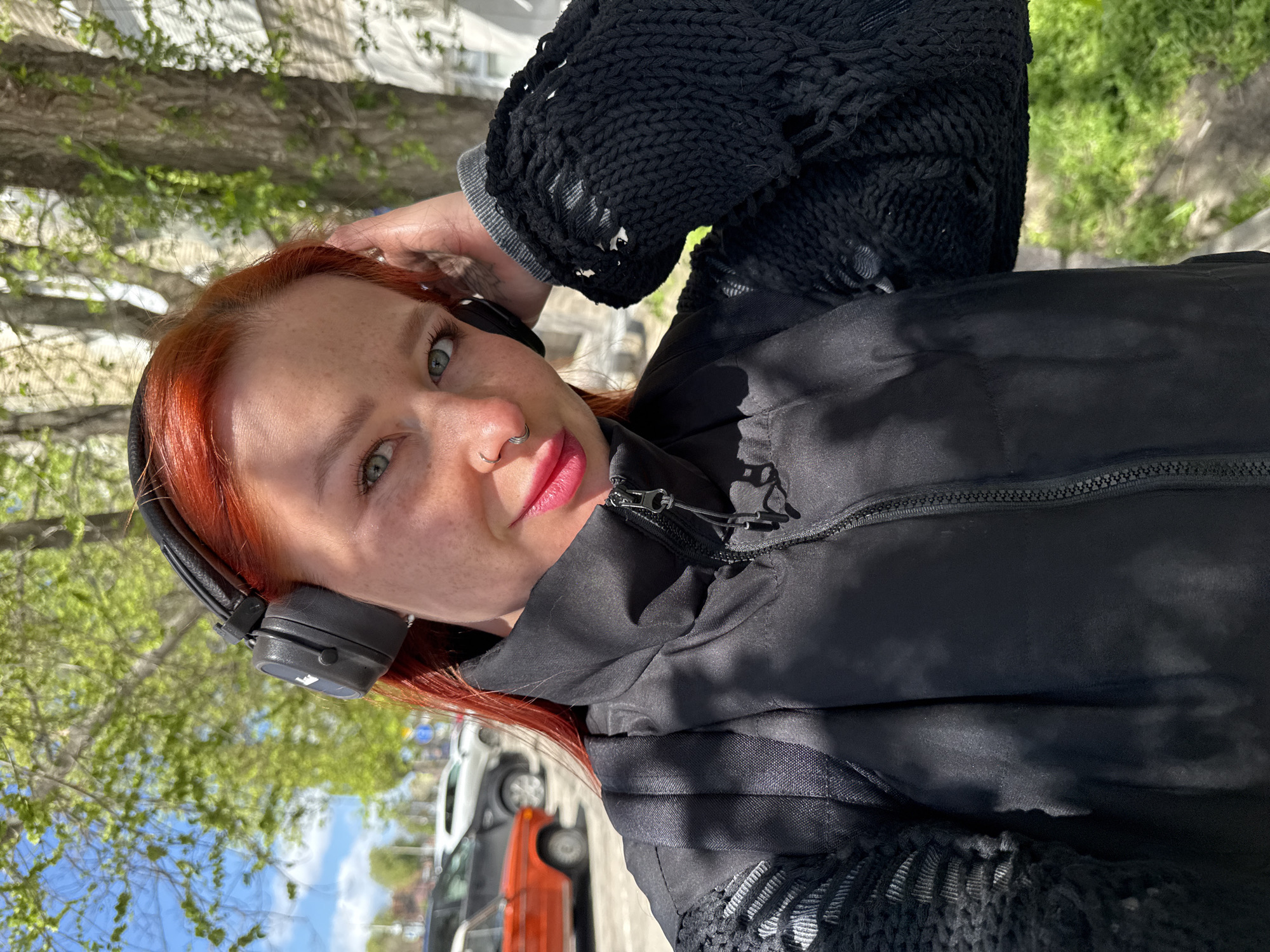 A young woman with red hair and freckled skin is posing for a selfie outdoors. She wears headphones and a black jacket with a textured knit sleeve. The background shows a city street with trees, parked cars, and buildings under a partly cloudy sky. While the location is likely in a city, there is insufficient information to confirm Rostov-on-Don, Russia.
