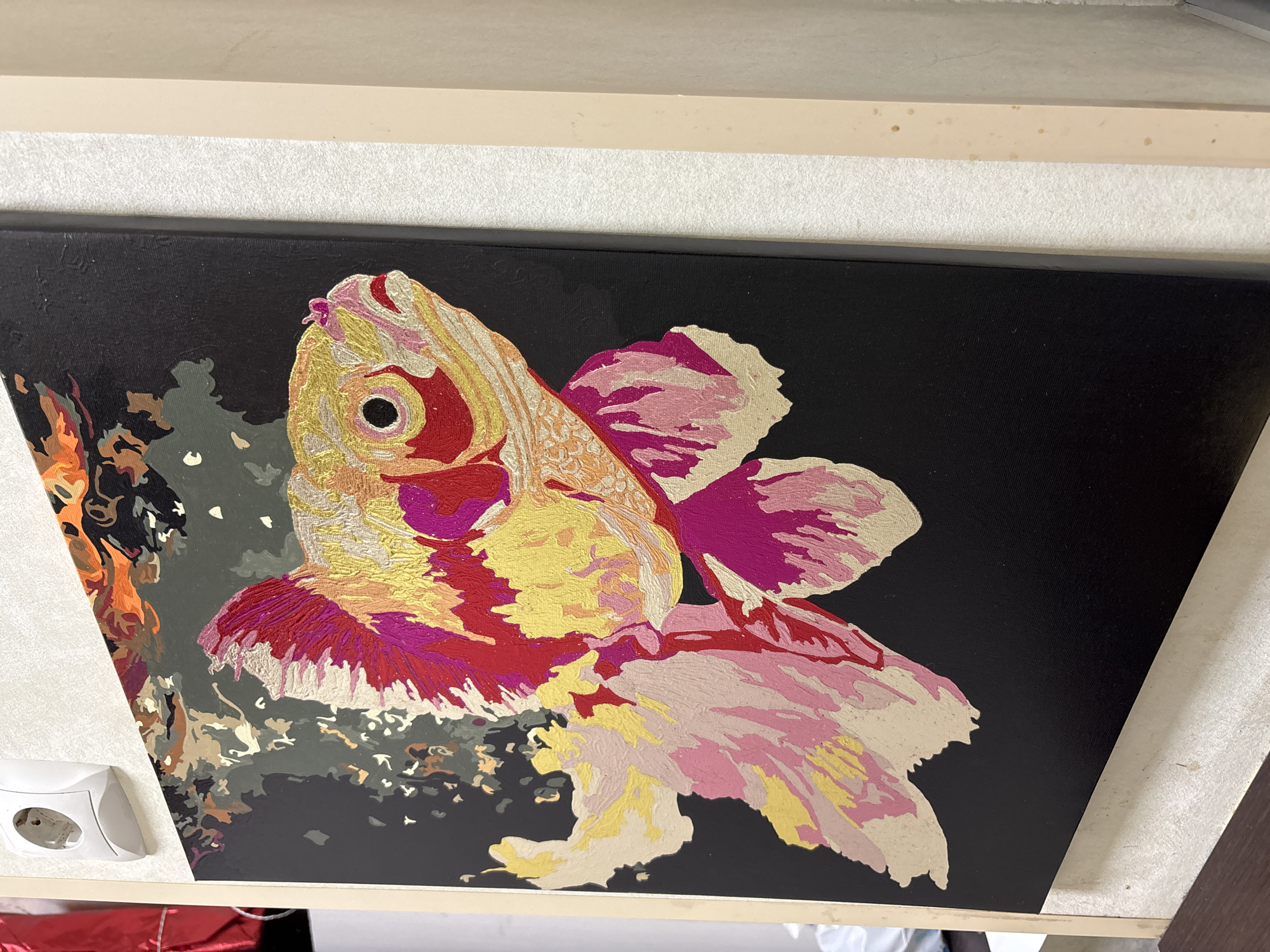 A painting of a goldfish is displayed on a wall. The fish is composed of various colors, including red, pink, gold, and beige. The background of the painting is black. The painting is framed and mounted on a wall in a room, which may be located in Vsevolozhsk, Russia. Other items are visible, including a wall outlet and a piece of furniture below the painting.