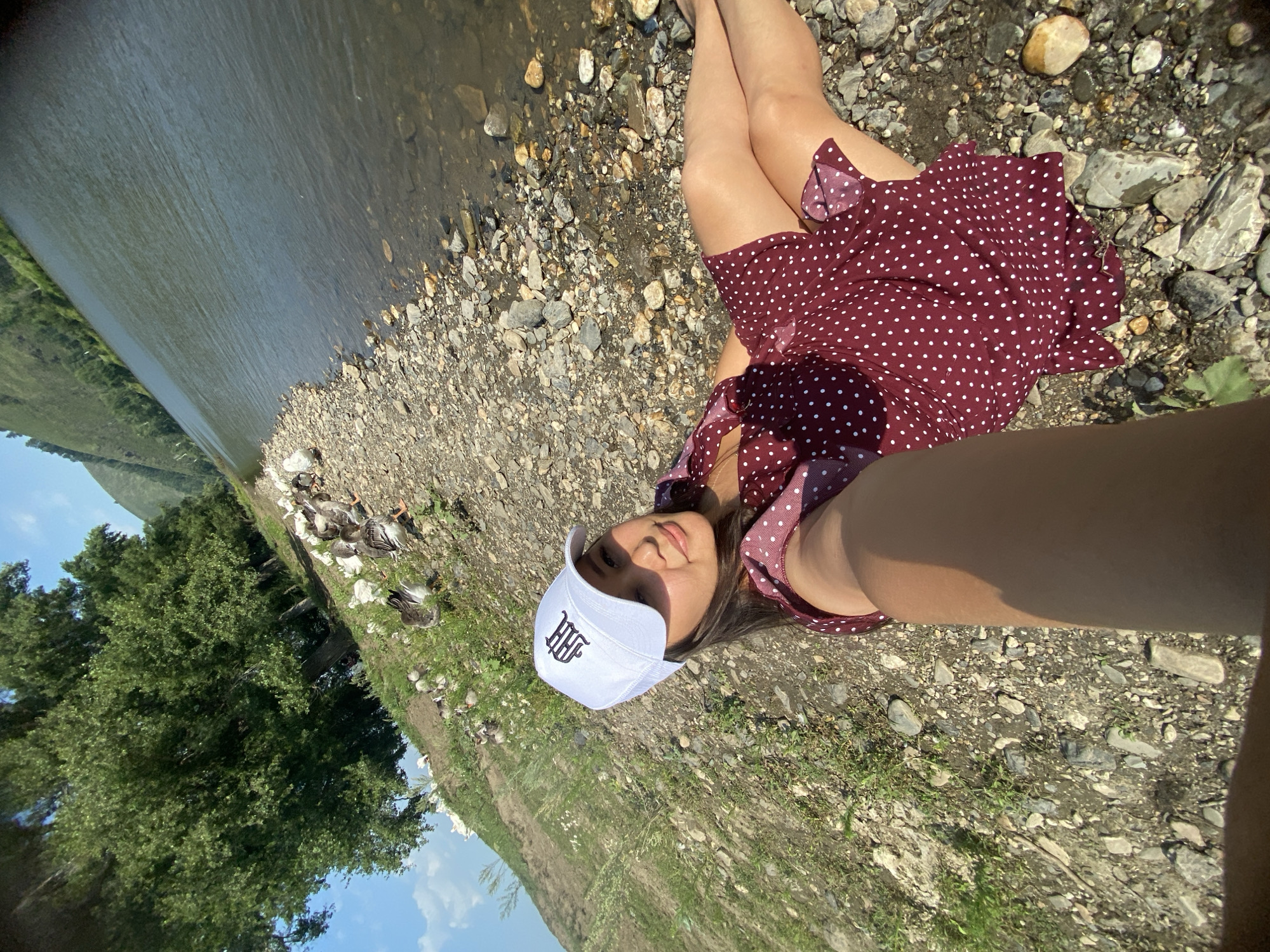 A young woman wearing a white baseball cap with an emblem and a maroon polka-dot dress is lying on a rocky shore next to a river. She is holding a phone in her right hand, taking a selfie. A flock of geese are visible in the background on the grassy bank near a large tree. The river flows to the right of the frame, and a tree-covered hillside rises on the opposite bank. The sky is blue with scattered clouds. The scene is consistent with a natural landscape, potentially near Maloarslangulovo, Russia, given the environmental cues.