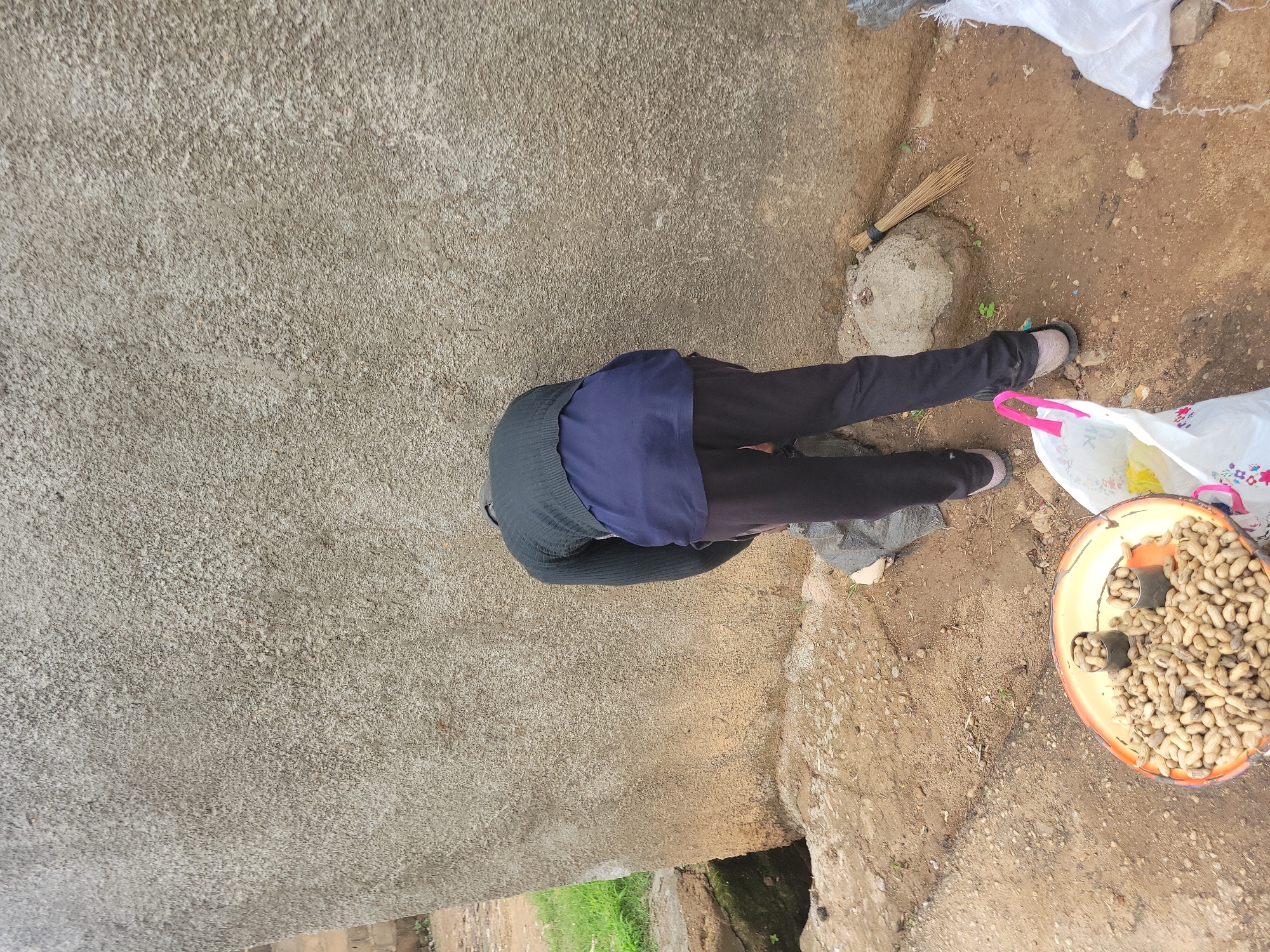 A person, viewed from behind, is bending over next to a textured concrete wall. They are wearing a dark long-sleeved sweater over a blue garment, dark pants, and sandals. In the foreground, to the left, is a shallow, orange-rimmed bowl filled with peanuts. To the right of the person, a broom leans against a rock near the wall. A white plastic bag is also visible on the ground to the right. The ground is a mixture of dirt and small stones, with some patches of dry grass. There is a dark opening in the ground to the left of the scene. The overall impression is of an outdoor, possibly informal market or residential area.