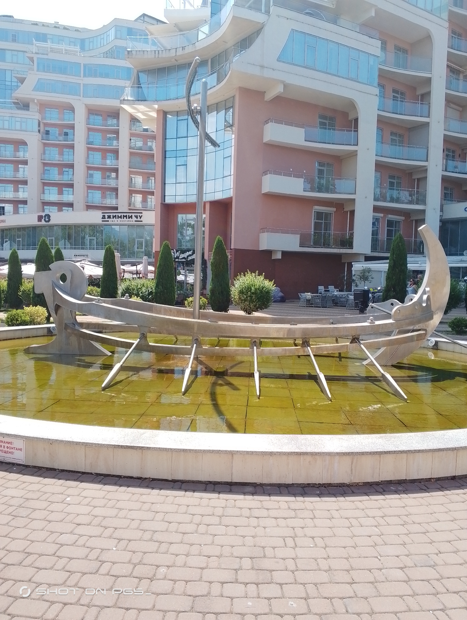 A contemporary fountain sculpture, resembling an abstract boat with oars, is the central subject. It is situated in a circular fountain basin filled with murky green water. The sculpture itself is metallic, with a sleek, modern design, featuring a curved hull and several pointed oar-like protrusions.
Behind the fountain, a modern building complex dominates the scene. The architecture is characterized by curved facades, numerous balconies with glass railings, and extensive use of glass windows, giving it a contemporary and somewhat opulent appearance. The building's exterior is a soft pink and cream color.
To the left of the building, a sign with Cyrillic text is visible. It reads "ДЖИММИ ЧУ" which translates to "JIMMY CHOO". Below this, in smaller text, it says "ЕДА И КОКТЕЙЛИ" meaning "FOOD AND COCKTAILS". Another sign with Cyrillic text is partially visible on the far left, reading "ВНИМАНИЕ!" which means "ATTENTION!".
The setting appears to be a public space, possibly a promenade or plaza, given the brick-paved ground surrounding the fountain. Several tall, slender trees are planted around the fountain and in front of the building. A few outdoor tables and chairs are visible in front of the building, suggesting a cafe or restaurant area. The lighting indicates it is daytime, with bright sunshine casting shadows. The overall atmosphere is one of a modern, sunny resort or urban environment. No people are clearly depicted in the foreground or midground, though there might be indistinct figures in the background near the building.