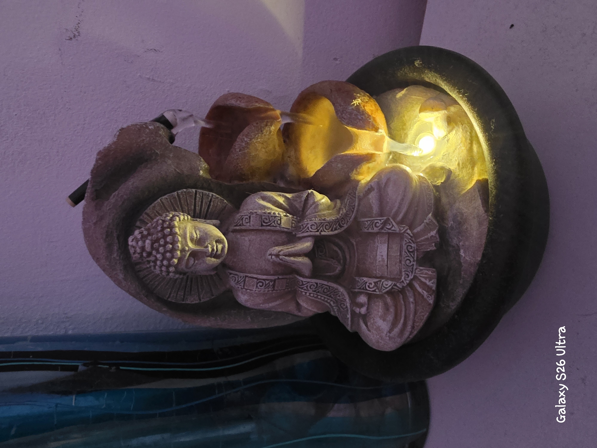 This image features a decorative tabletop fountain depicting a serene Buddha statue. The fountain is made of a light-colored, textured material, possibly resin or ceramic, designed to resemble stone. The Buddha is seated in a meditative pose with its eyes closed and a gentle smile. Water appears to be flowing from behind the Buddha's head and into the basin. The water is illuminated by a warm yellow light, creating a tranquil ambiance. The fountain is situated against a textured, purple-tinted wall. Below the fountain, there is a reflective surface with wavy patterns in shades of blue and black. The scene is indoors, likely in a home or a meditation space, given the decorative nature of the object. The lighting suggests it could be evening or a dimly lit room. There is no clear indication of the weather or time of day outside the immediate setting. The overall mood evoked is one of peace and calm.
Visible text in the image reads "Galaxy S26 Ultra," which is likely a watermark from the device used to capture the photograph. The location context provided is Plovdiv, Bulgaria.