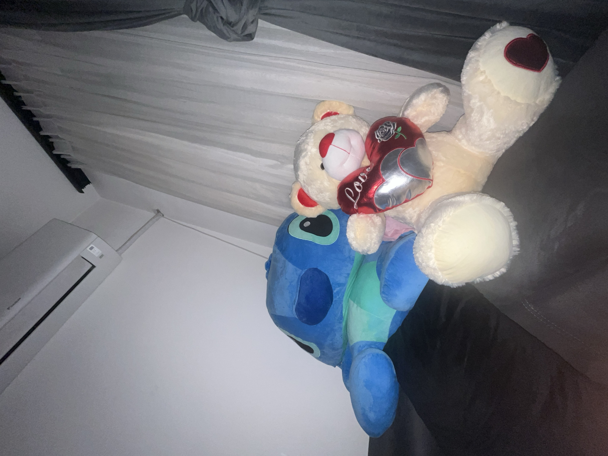 This indoor media file, likely captured in Plovdiv, Bulgaria, displays two large plush toys: a cream-colored teddy bear and a blue Stitch character, resting on a dark surface, possibly a bed or couch. The image is rotated, making the wall with the air conditioning unit appear on the left and the ceiling/curtains overhead.
The cream-colored teddy bear is positioned on top, embracing a red and silver heart-shaped pillow that clearly displays the word "Love" in white script. The teddy bear features a red heart detail on its nose and inside one ear, and a larger, darker red heart on its right foot. Beneath the teddy bear, the blue Stitch plush toy is partially visible, showing its characteristic blue fur, large ears, and black eyes.
In the background, a white air conditioning unit, faintly showing the "Daikin" brand, is mounted on a light-colored wall to the left. Above the toys, light, sheer curtains are draped, with a darker grey fabric gathered at the top, suggesting window coverings or a canopy. The overall setting appears to be a cozy personal space, such as a bedroom or living room. No people are present, and no specific activity is taking place; it's a still arrangement of beloved items. The lighting indicates an indoor environment, but the time of day is not discernible. The plush toys, especially the heart-holding teddy bear, evoke feelings of warmth and affection.