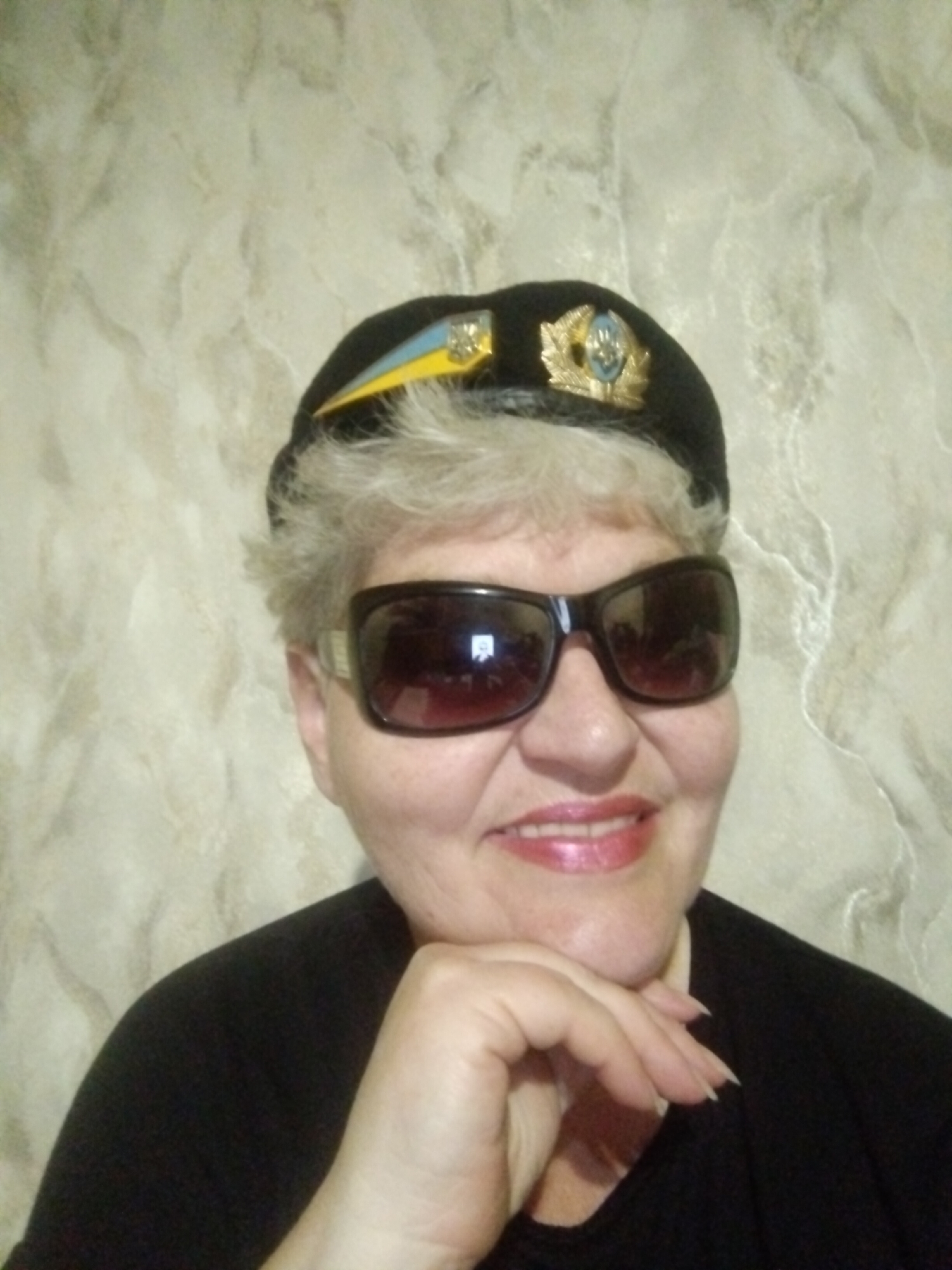 An older woman wearing a black beret with a yellow and blue emblem and gold accents, dark sunglasses, and a black top, poses for a selfie indoors. She has short, light-colored hair and is smiling with her chin resting on her hand. The background is a textured beige wall. The image was taken in Horishni Plavni, Ukraine.
