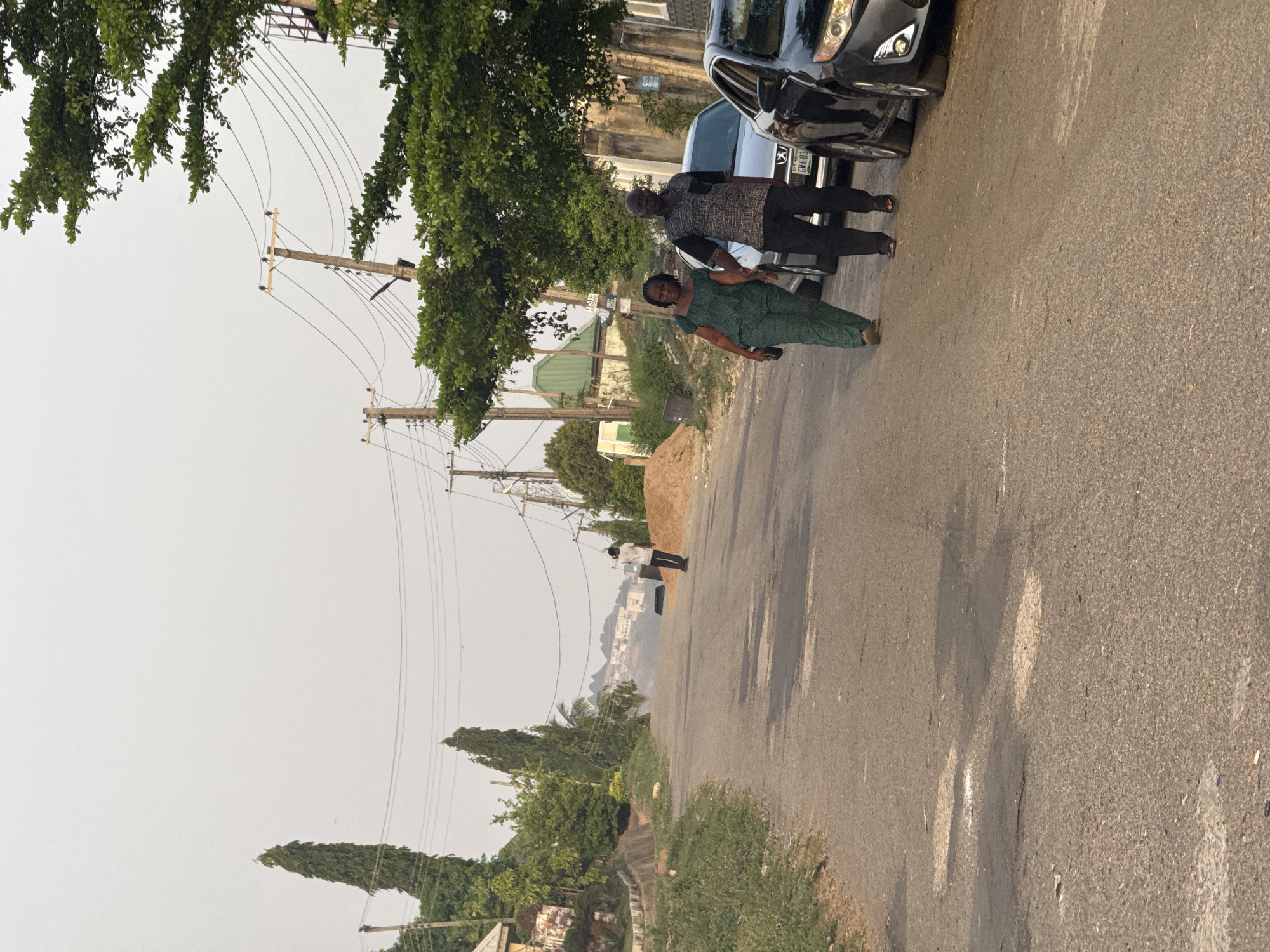 An outdoor street scene in Abuja, Nigeria, captured during what appears to be late afternoon or early morning, indicated by the quality of light and shadows. The sky is bright and clear. The image shows a paved road, possibly asphalt, with two adults standing in the foreground on the right side. A woman wearing a green patterned outfit is holding a phone in her hand, facing slightly towards the camera. Next to her, a man in a patterned dark shirt and black trousers stands, looking towards the camera. Both individuals are standing beside at least two parked cars. The front of a black car with a partially visible license plate reading "GWA 03" is prominent.
Further down the road, another person in a white shirt and dark trousers can be seen walking away from the camera. The left side of the road features several wooden utility poles with numerous overhead wires, interspersed with lush green trees and foliage. In the middle distance, there are some buildings and a pile of sand or dirt. The background reveals a panorama of distant hills or mountains, with residential structures visible on their slopes. The overall impression is a typical street view in a residential or suburban area. No strong emotions are discernible from the people.