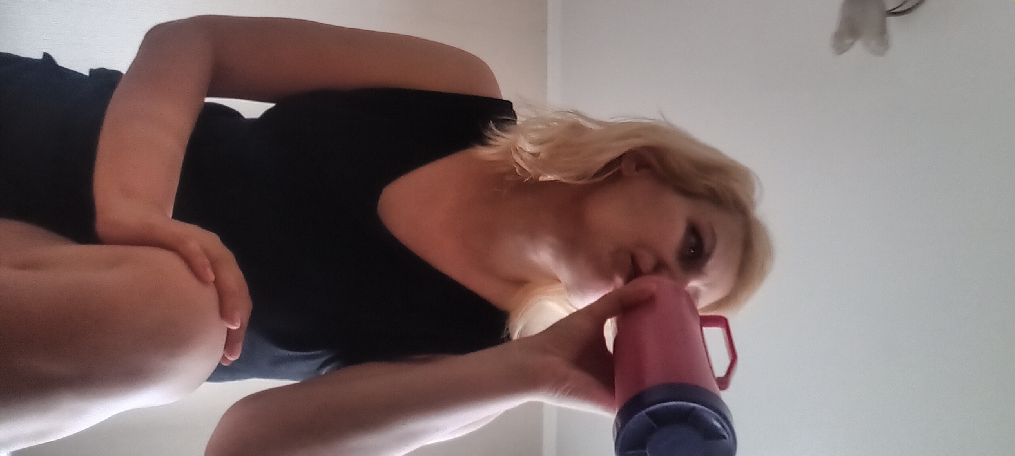 A blonde woman in a black dress is holding a pink and blue object to her face, appearing to drink from it. Her left hand is resting on her knee, and her right arm is raised. The background is a light-colored wall and ceiling, with a light fixture visible in the upper left corner. The setting appears to be indoors, likely a residential space. There is no visible information to suggest the location is in Kyiv, Ukraine.