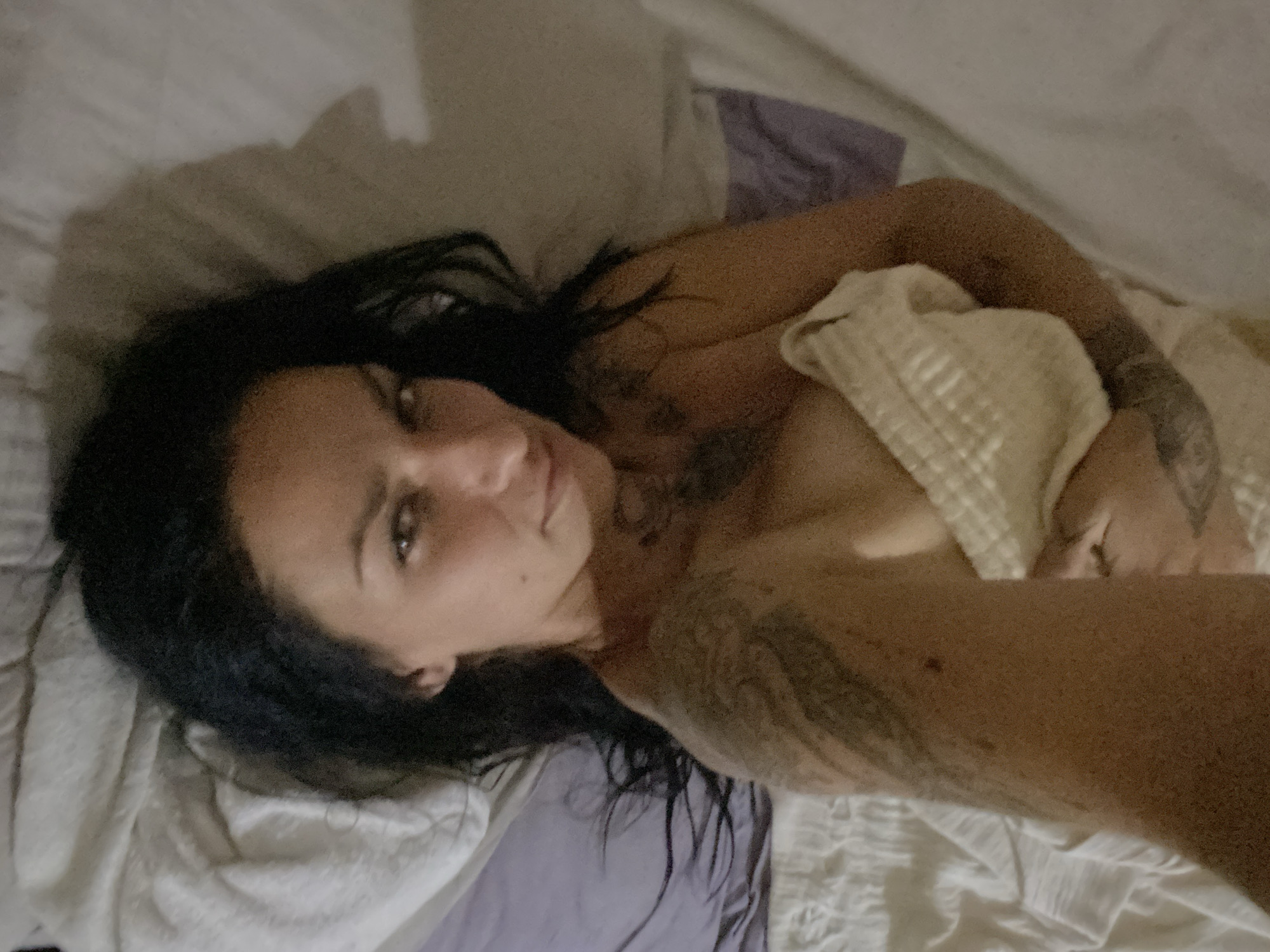 This image depicts a single adult female in an indoor bedroom setting, specifically in Mũi Né, Vietnam, as designated by the user. The woman is positioned lying supine on a bed adorned with white or light-colored sheets and a pillow. Her dark, wavy hair is spread across the pillow. She possesses dark eyes that are directed towards the camera, accompanied by a subtle facial expression. Distinct tattoos are visible on her left upper arm, shoulder, and potentially on her chest area. Her upper torso is partially concealed by a light-colored, textured fabric, likely a towel or blanket, which she holds across her chest. Her right arm is visible, extending towards the bottom left of the frame, suggesting she is holding the camera for a selfie. The lighting within the scene is subdued, and a prominent shadow, consistent with a head profile, is cast on the pillow to the right of her head, indicating a specific light source. A bunched purple sheet or fabric is present on the bed to the left of the woman.