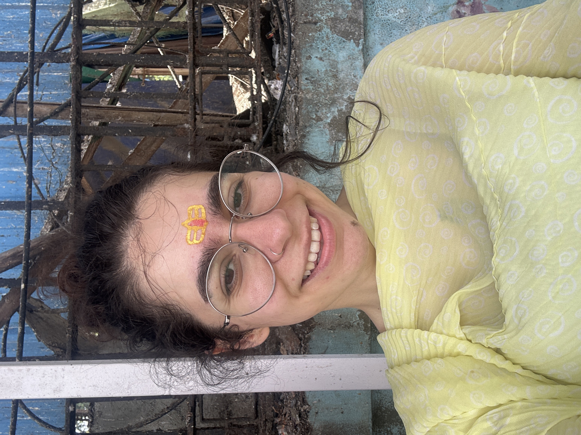 A young woman with dark curly hair pulled up in a messy bun smiles at the camera. She wears round, silver-rimmed glasses and a small hoop nose ring. A red and yellow religious marking is visible on her forehead, between her eyebrows. She is draped in a light yellow, patterned scarf. Behind her, a weathered blue wall is visible, partially obscured by a dark, rusted metal grate or fence. The scene appears to be outdoors, with elements suggesting an urban environment in Mumbai, India.