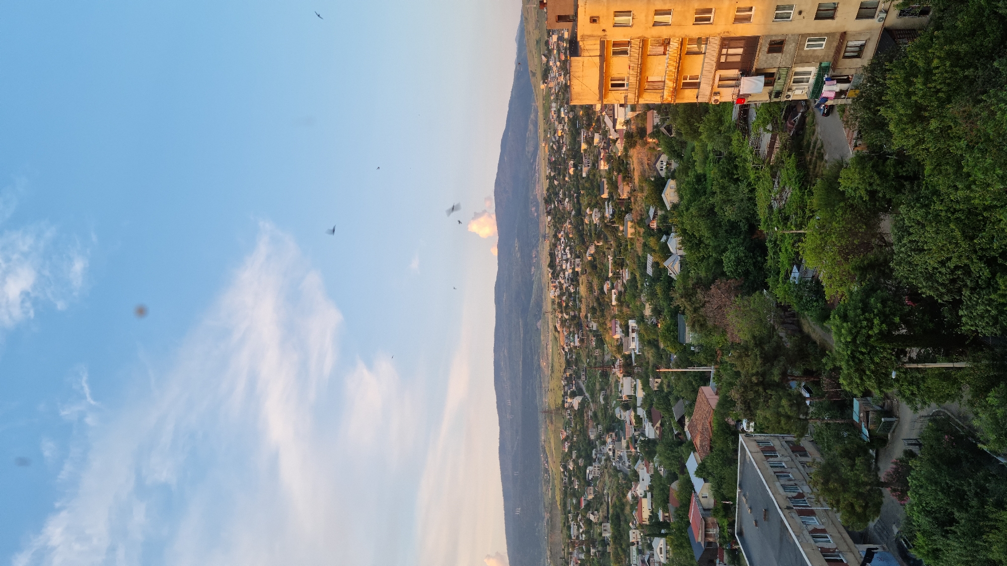 The image shows an aerial view of a residential area in Tbilisi, Georgia, likely in the early evening as indicated by the lighting. In the foreground, on the right, is a multi-story apartment building with a weathered, light brown facade. Laundry is hanging from balconies on the middle floors. To the left of the apartment building, there are numerous trees and smaller houses with red and white roofs, characteristic of many Tbilisi neighborhoods. A longer, grey apartment or institutional building with visible windows is prominent in the lower left corner of the image. The middle ground is a dense expanse of green trees interspersed with houses. In the background, a wide, rolling mountain range forms the horizon, with some power lines visible on its slopes. The sky is a clear blue with scattered white clouds and several birds are visible in flight. The overall impression is of a densely populated urban landscape blending with a natural mountainous backdrop.