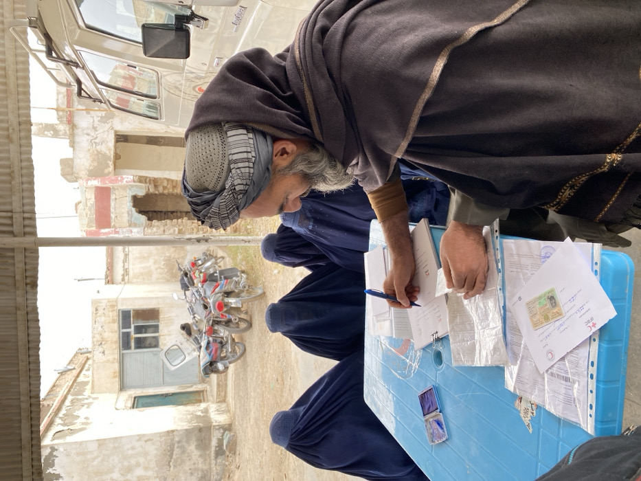 An elderly man with a gray beard and turban fills out paperwork at a blue table. Three women in blue burkas stand in line. Motorcycles are parked near a building with peeling paint in the background. Documents, some with photos, are scattered on the table. A blue inkpad is also visible. The scene appears to be taking place outdoors in a somewhat rural or impoverished area.