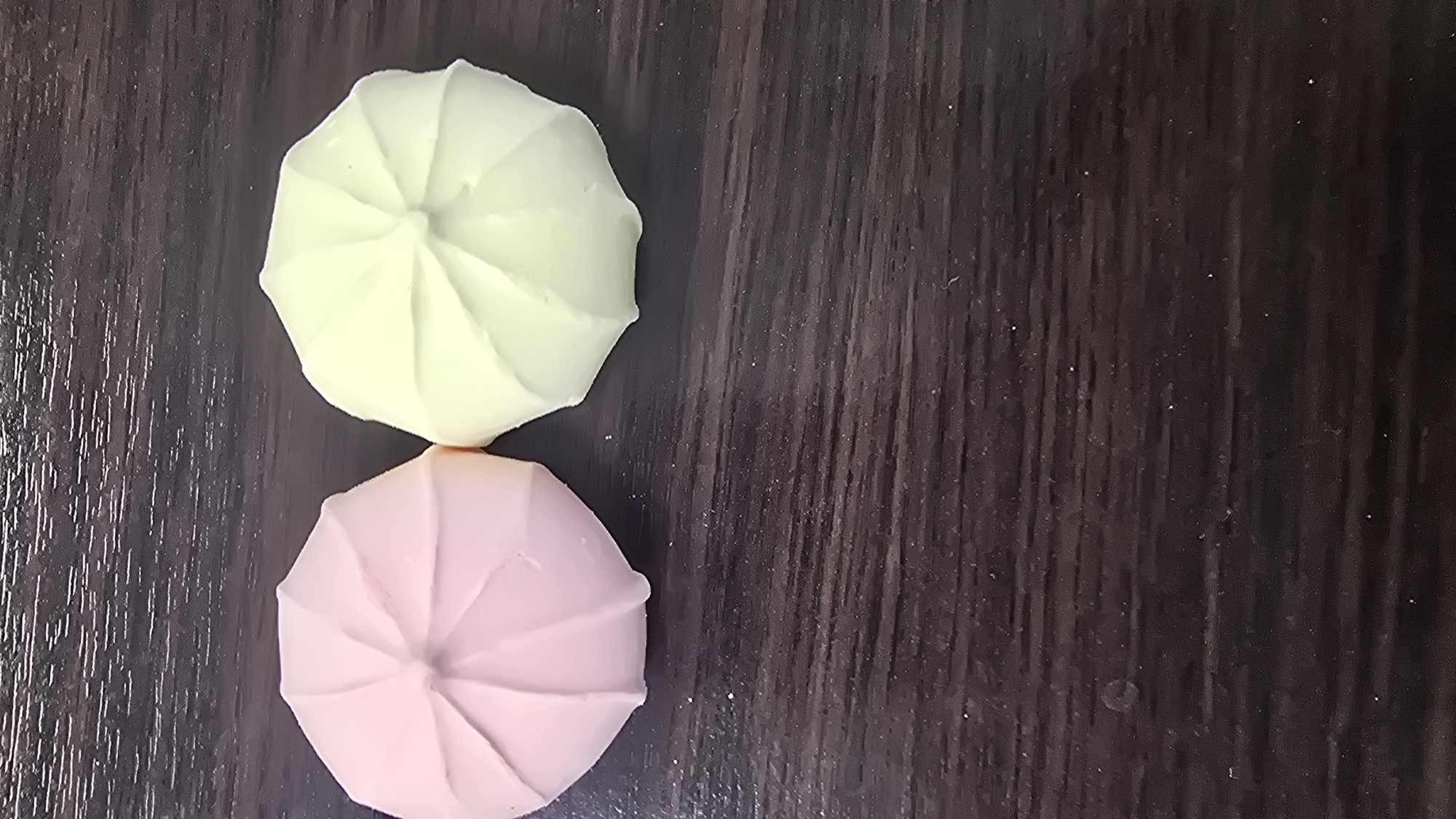 A top-down view displays two identical, dome-shaped confections, one pink and one pale yellow, resting on a dark brown wooden surface. Both confections exhibit a symmetrical radial, segmented pattern on their upper surface, resembling a closed umbrella or a fluted design. The pink confection is positioned to the left of the yellow one, with their sides nearly touching. The dark brown surface, likely wood, displays a prominent horizontal grain pattern and scattered tiny white specks, consistent with dust or fine powder. The lighting is even, creating soft shadows beneath the objects. This scene is located in the city of Kazan, Russia.