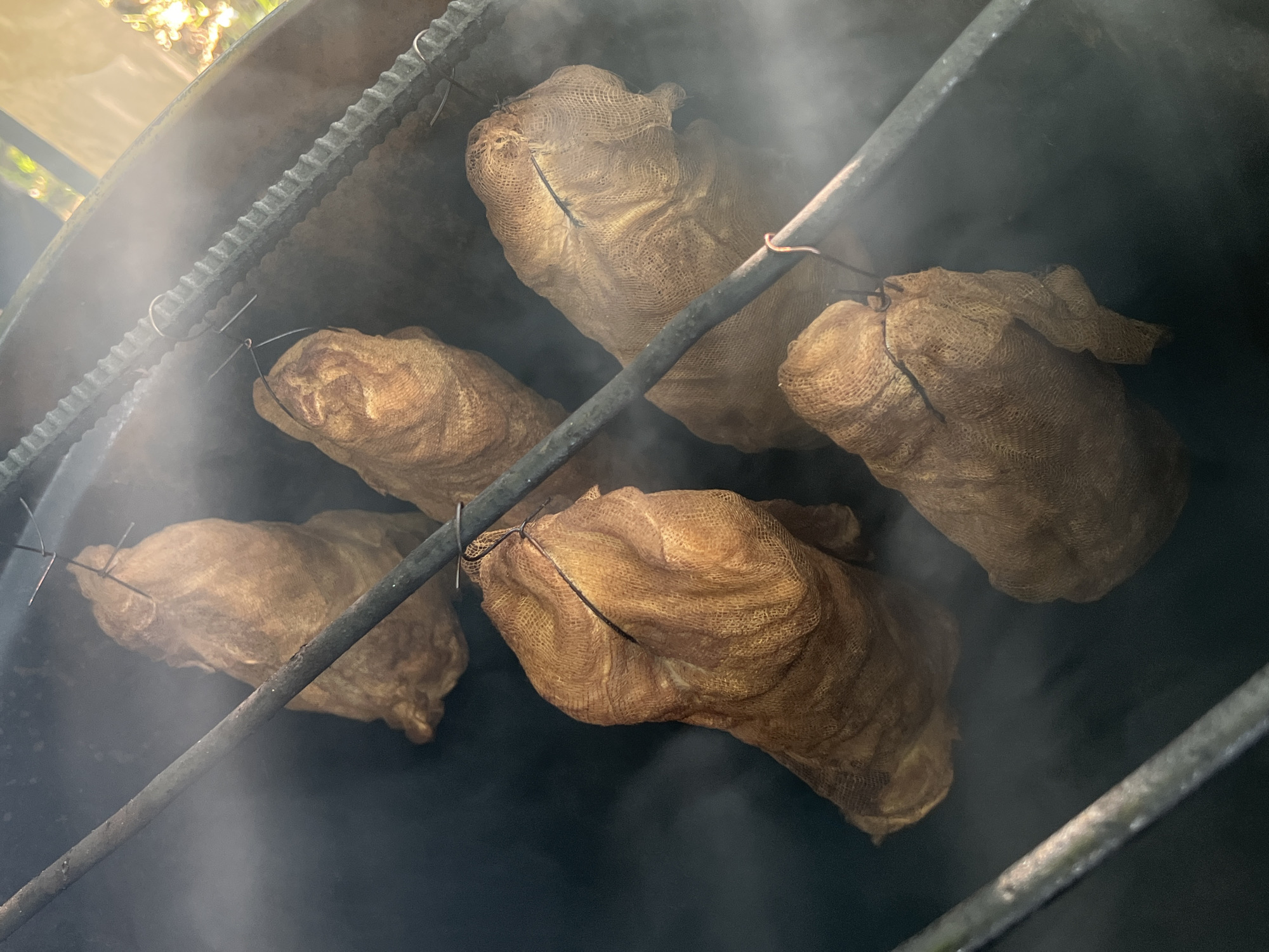 Five netted parcels, likely food items, are suspended by metal hooks and rods within a dark, smoky enclosure, possibly a smoker or grill. The parcels are light brown and appear to be made of a burlap-like material, with visible mesh texture. They hang at varying heights and angles. Wisps of smoke or steam drift around the parcels and obscure parts of the background. The enclosure itself is dark, with some metal framing visible. The overall scene suggests a food preparation or smoking process. There is no indication of a specific location in Lublin, Poland.