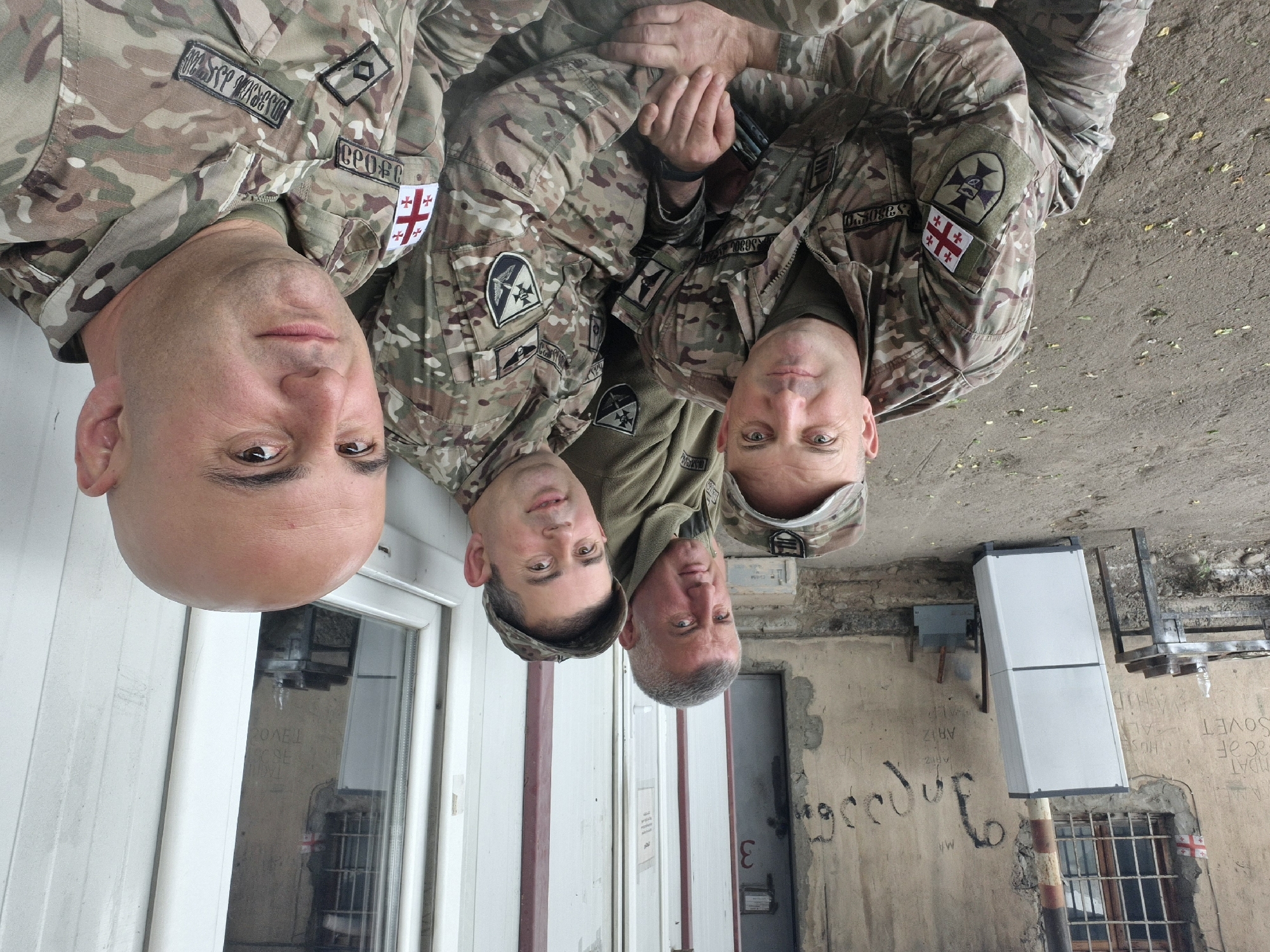 Four uniformed individuals, likely military personnel, are positioned outside a building with a distressed exterior in Tbilisi, Georgia. All are wearing Georgian military camouflage uniforms with the Georgian flag patch. The bald person is in the foreground near a white door and window. Behind them are three additional uniformed individuals, a white painted building with graffiti. A Georgian flag is seen in a nearby window. The building appears old and neglected, and the presence of Georgian flag patches suggests the subjects are likely part of the Georgian military.