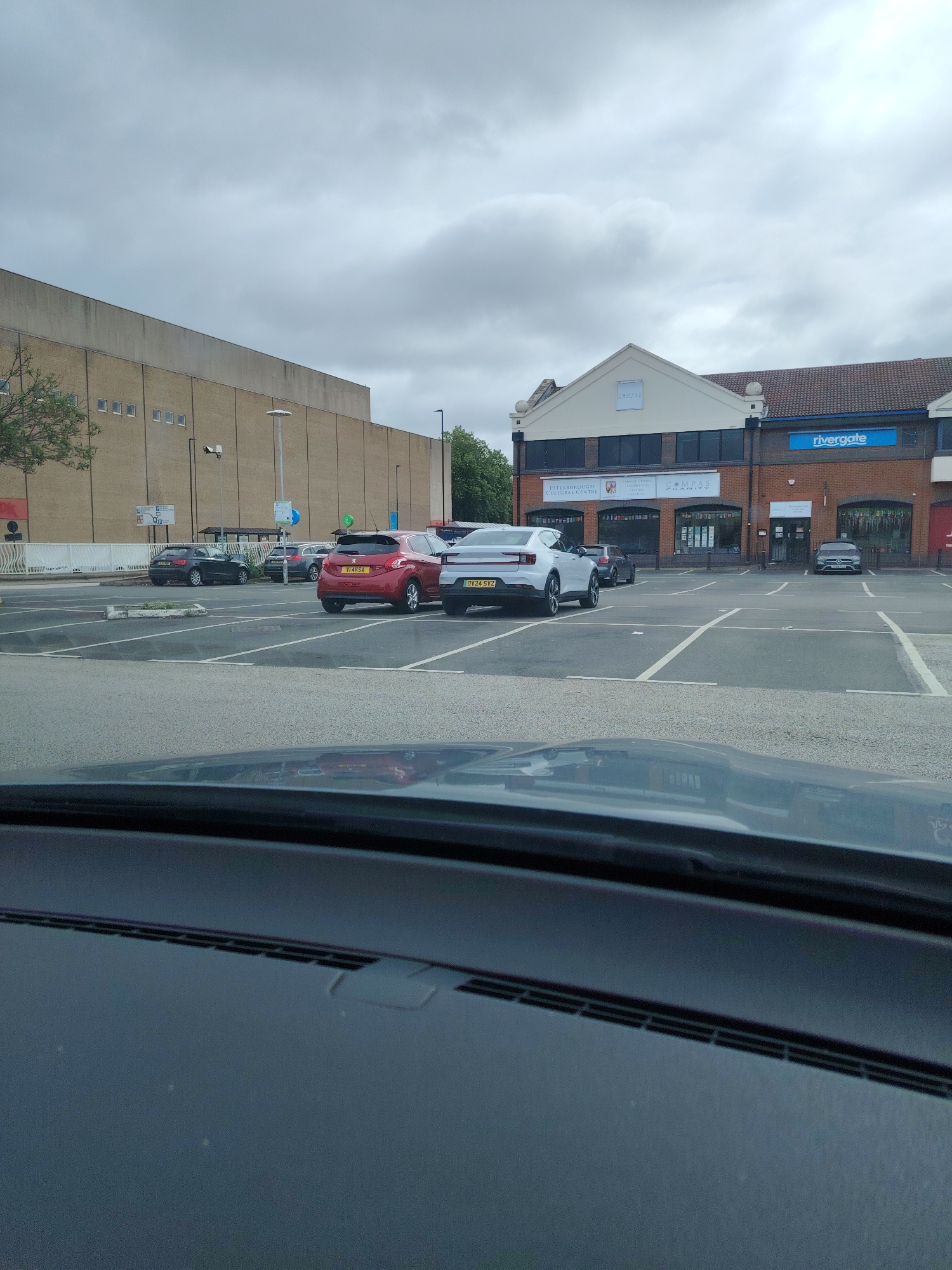 The image displays a parking area in the city of Peterborough, United Kingdom, specifically adjacent to the Rivergate Shopping Centre and the Peterborough Cultural Centre. The perspective is from inside a vehicle, with the dashboard and windshield visible in the immediate foreground.
In the midground, a paved parking lot is present, marked with white lines indicating parking spaces, many of which are empty. Several vehicles are parked or visible within the lot. Prominently featured are a red Peugeot 208 hatchback (license plate VI14 KSA) parked perpendicularly to the camera's view, and a white Polestar 2 electric car (license plate OV24 SVZ) parked to its left, angled slightly towards the camera. A dark BMW SUV is visible further back on the left side of the parking lot, and other vehicles are partially obscured. Parking signs, including those indicating disabled parking, are visible on light poles within the lot.
In the background, two distinct architectural structures are present under an overcast sky. To the left, a large, multi-story building with a plain, light brown/beige facade and rows of small, rectangular windows dominates the horizon. To the right, a two-story commercial or institutional building with a pitched roof section is visible. This building features prominent signage: a blue "rivergate" sign on the right, a white "compass" logo higher up in the center, and below it, storefronts displaying signs for "PETERBOROUGH CULTURAL CENTRE", "COMPAS CHARITIES", and an "Age UK" logo. The upper floor of this building features dark-tinted windows. No individuals are visible within the frame.