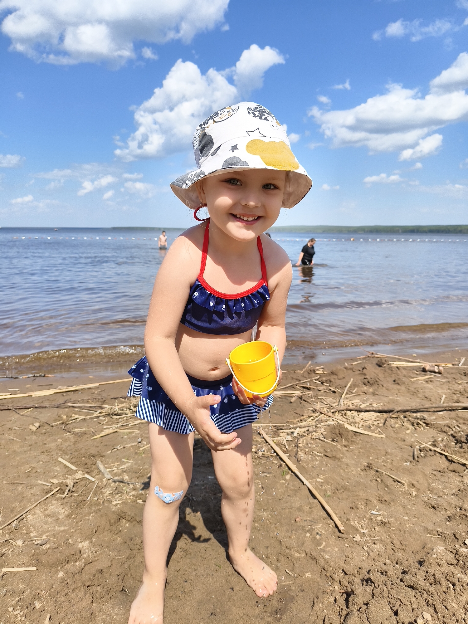 A young child stands on a sandy beach near a body of water, possibly a lake or river. The child is wearing a navy blue and white striped swimsuit, a white sun hat with a patterned design, and has a bandage on her left knee. She is holding a yellow bucket in her hands. The background includes blue sky with white clouds, and other people in the water. The foreground shows sand mixed with small pieces of wood debris. The city and exact location are Unknown, Unknown.