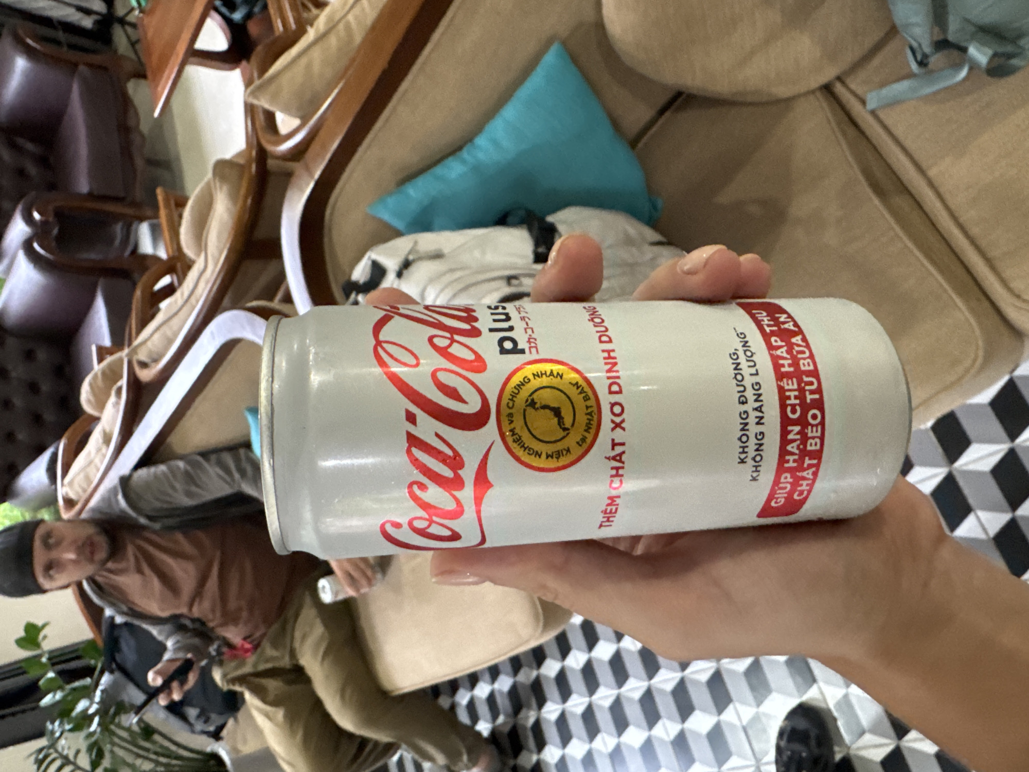 The image depicts an indoor scene, likely a cafe or living room, characterized by a striking black and white geometric tiled floor. In the foreground, a hand holds a frosted white can of Coca-Cola Plus. The can's surface suggests it is cold.
In the blurred background, another person is seated on a light brown sofa or armchair. This individual wears a brown long-sleeved shirt and a dark beanie, holding a pen-like object. Surrounding them are various pieces of wooden furniture, including chairs with light cushions and a sofa with a blue throw pillow. A green potted plant is partially visible on the left.
The can of Coca-Cola Plus displays prominent text in both Vietnamese and Japanese. Key Vietnamese phrases include: "THÊM CHẤT XƠ DINH DƯỠNG" (Added Dietary Fiber), "KHÔNG ĐƯỜNG, KHÔNG NĂNG LƯỢNG" (No Sugar, No Calories), and "GIÚP HẠN CHẾ HẤP THU CHẤT BÉO TỪ BỮA ĂN" (Helps Limit Fat Absorption from Meals). A circular seal indicates "KIỂM NGHIỆM và CHỨNG NHẬN tại NHẬT BẢN" (Tested and Certified in Japan). The Japanese text "コカ・コーラプラス" (Coca-Cola Plus) is also visible. The setting, with its specific can text, is consistent with Đà Nẵng, Vietnam. The scene captures a casual, relaxed moment indoors, under ambient lighting.