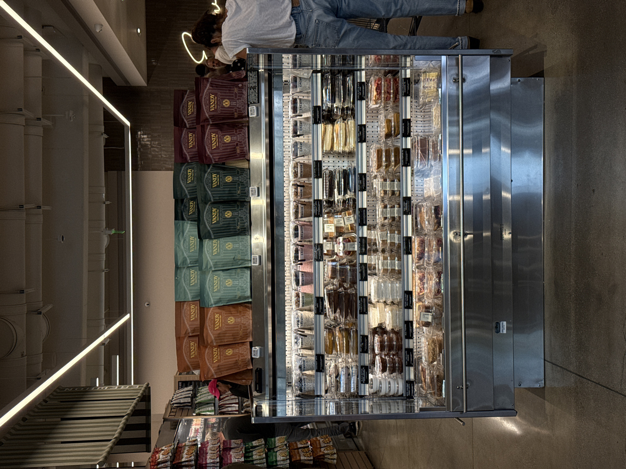 Here is a forensic description of the image:
The image shows the interior of a grocery store. A refrigerated display case is prominently featured, stocked with items like packaged foods, including “VANDY” brand products. A person wearing jeans and a light-colored shirt is standing near the display case, their upper body visible. The refrigerated display case is also stocked with REWHON and WHON-labeled products. The lighting inside the store is bright, with visible light fixtures on the ceiling. The overall setting suggests a retail environment, specifically a grocery store. The location, Beverly Hills, United States of America, is not discernible from the image alone.