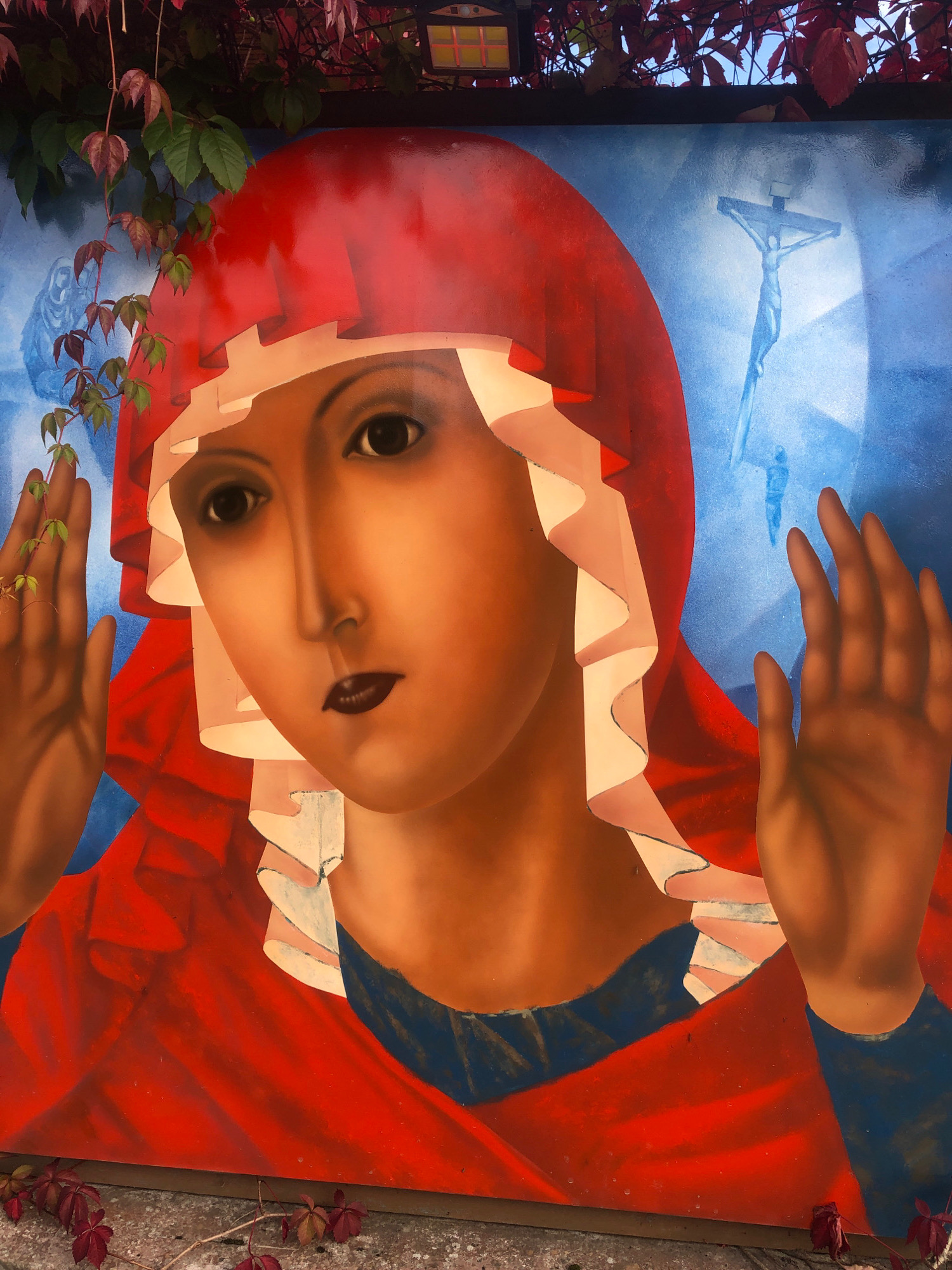 A large mural painting depicts a woman with dark hair and makeup, wearing a red headscarf and a red garment. She has her hands raised, palms outward. In the background, a blue sky with a faint outline of Jesus on the cross is visible. The painting is outdoors, with green and red leaves from a vine encroaching on the top and bottom edges. Some of these leaves are also on the ground in front of the mural. The location is identified as Intourist SNT, Russia.