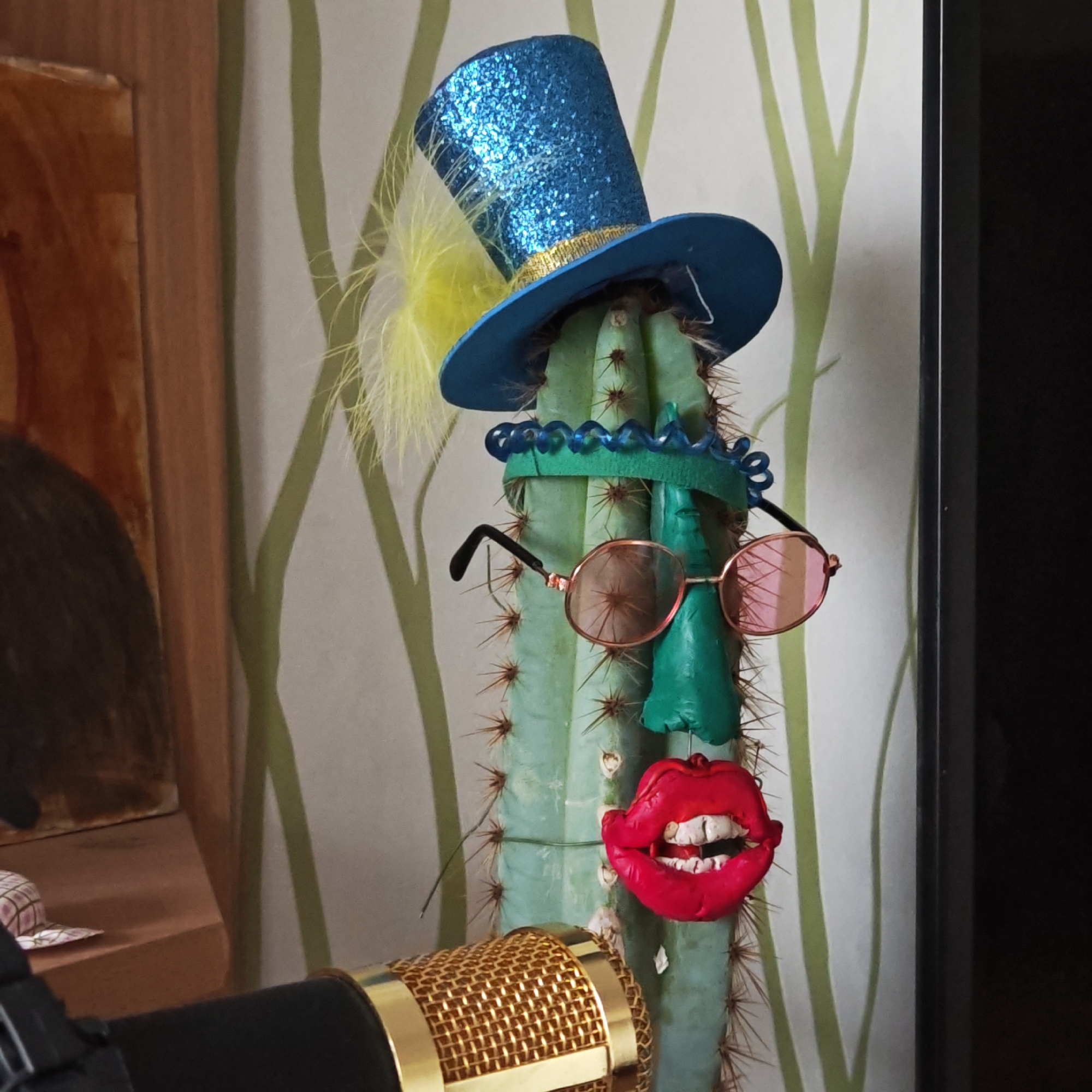 A medium shot features a cactus adorned with costume props, set against a wallpapered background with vertical green stripes. The cactus is decorated with a blue glittery top hat with a yellow feather, a blue beaded headband, rose-tinted sunglasses, a green nose, and red lips with white teeth. The lower left corner of the image includes a portion of a gold-colored microphone. The scene is indoors, and an indiscernible object appears in the background, partially obscured. The location and city of Ketoshi, Russia, cannot be definitively confirmed based solely on the given image.
