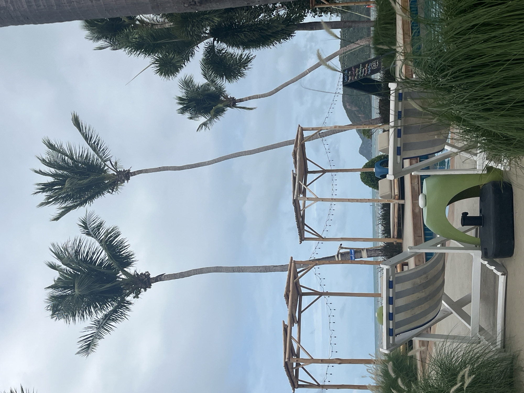 This image depicts an outdoor seating area at what appears to be a beachfront resort or restaurant on the island of Ko Pha-ngan, Thailand. In the foreground, two striped lounge chairs face a swimming pool and the ocean beyond. A green, curved stool is positioned between the chairs, and a black umbrella base is visible on the sand. Several palm trees are scattered throughout the scene, with their fronds reaching towards a cloudy, overcast sky. Two open-air wooden structures, possibly gazebos or shaded seating areas, are situated behind the lounge chairs, adorned with string lights. In the distance, lush green hills rise from the coastline, partially obscured by the trees. A blackboard sign is propped up near the pool, displaying what looks like a menu or event information. The overall atmosphere is relaxed and tropical, with the overcast weather suggesting a possible break in the sun.