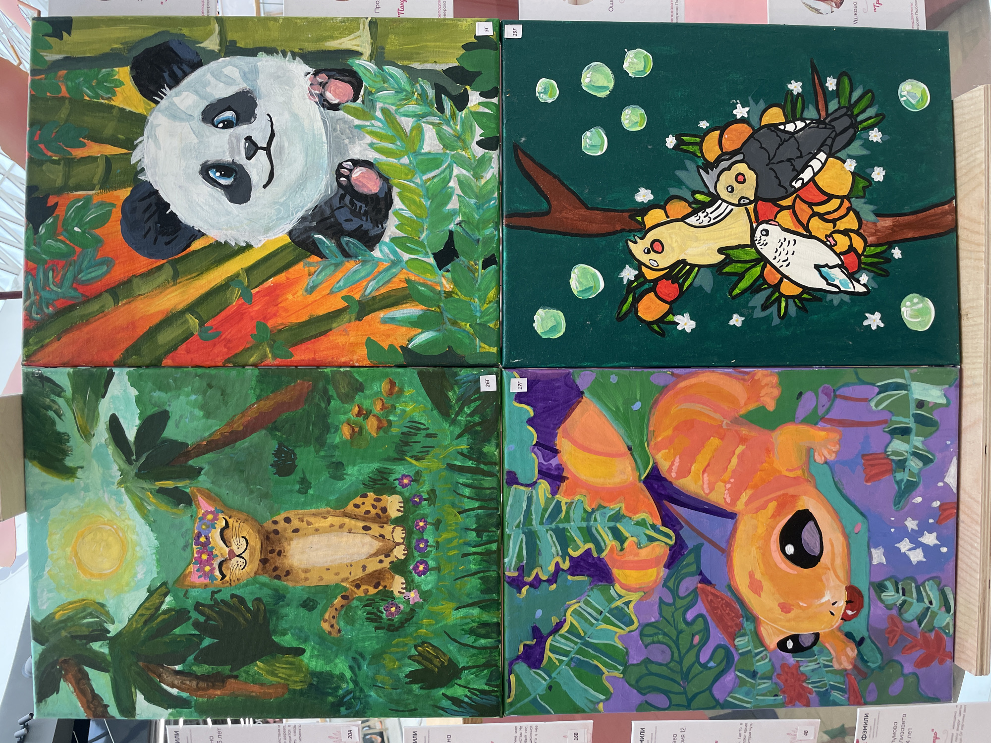 The image is a top-down view of four colorful paintings displayed in a grid. Each painting features a cartoonish animal. The top-left painting depicts a panda lying on its back amongst bamboo stalks and green leaves. The top-right painting shows three stylized birds perched on a branch with colorful fruits and small white flowers against a dark green background with some bubbly green circles. The bottom-left painting features a leopard lying down in a jungle-like setting with palm trees and a bright yellow sun. The bottom-right painting displays an orange squirrel-like creature with large black eyes, surrounded by vibrant green and purple foliage and some small white stars. The paintings appear to be child-made art, possibly from an exhibition or school project, as indicated by the numbered labels (e.g., "26Г", "17г", "29Г", "5г", "48", "20A") and visible text in Russian which includes names of teachers ("преподаватель Верова Любов") and children's names and ages, suggesting a local context like "Perm, Russia." The overall mood is cheerful and whimsical due to the bright colors and playful animal depictions. There are no people clearly visible in the paintings themselves.