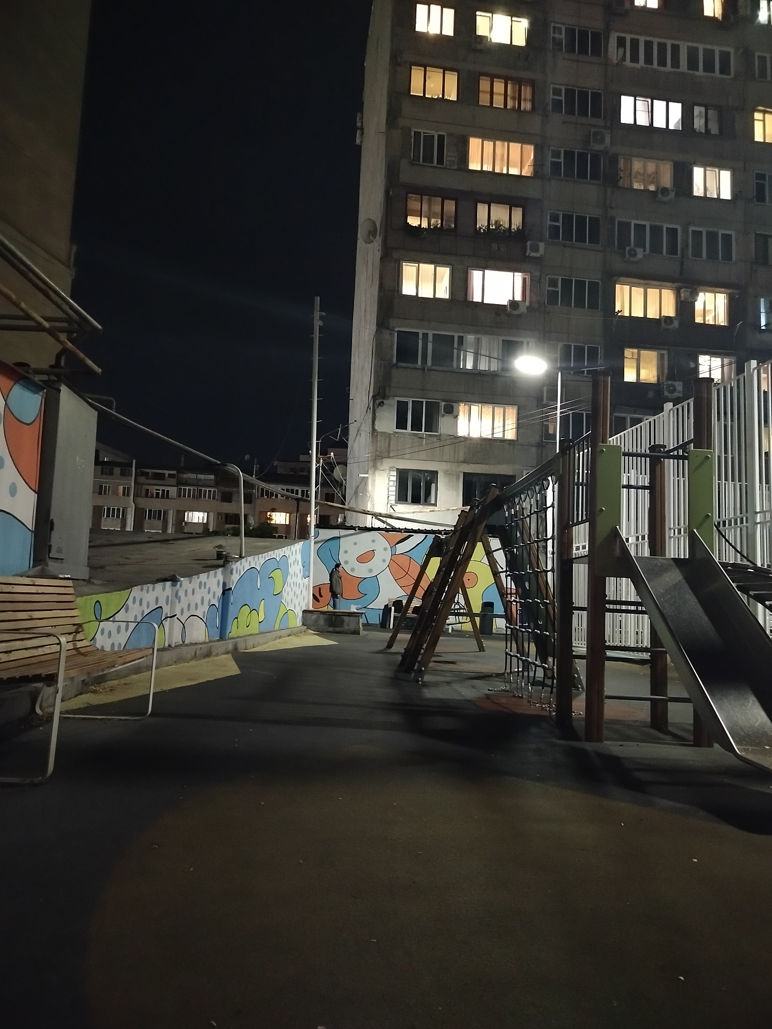 A playground at night in Yerevan, Armenia. The playground has a slide and other equipment. A colorful mural is painted on a wall. In the background, there is a large apartment building with many windows lit up. A streetlight illuminates the scene. The ground is paved with a dark surface. A bench sits beside the wall.
