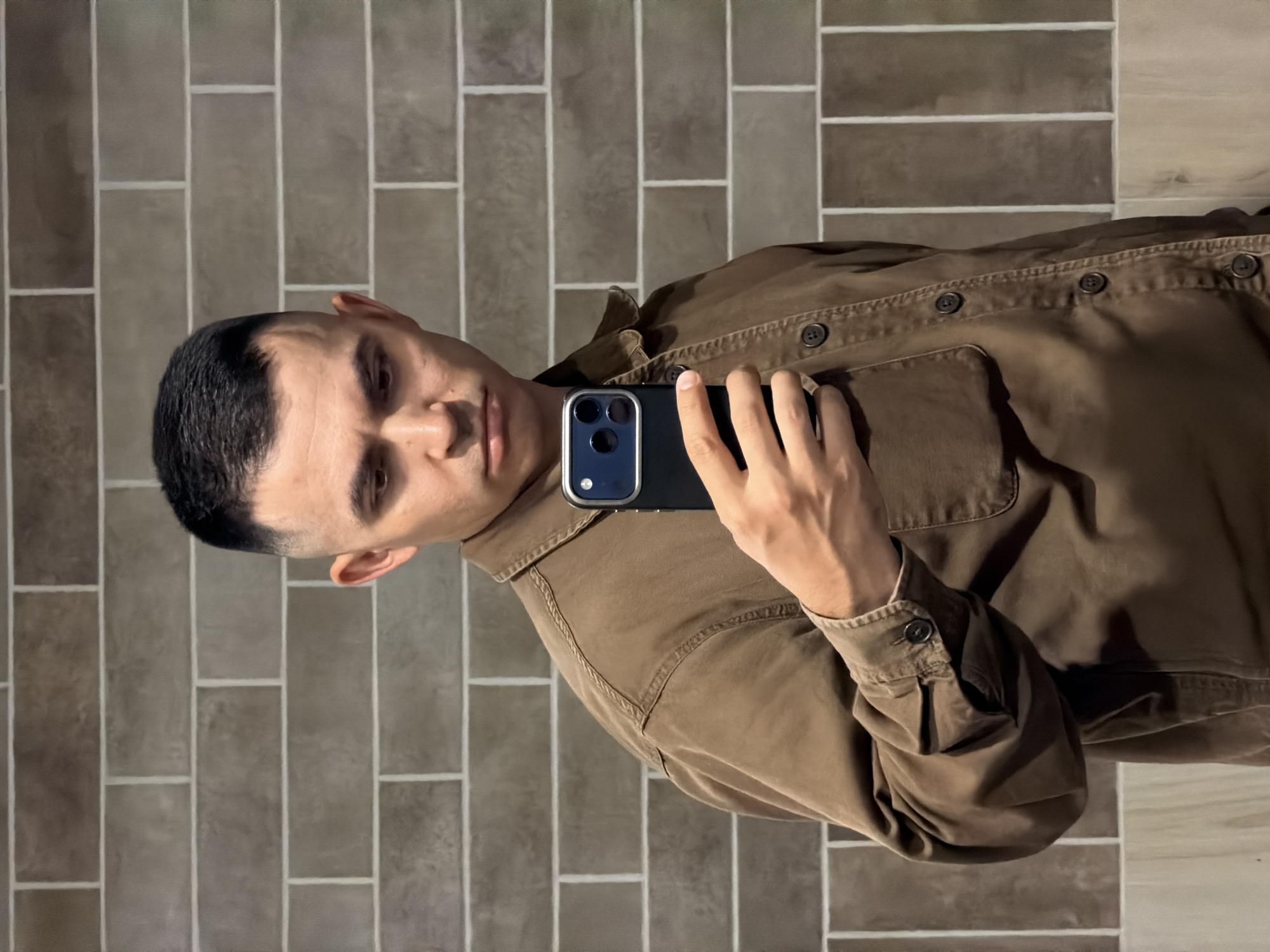A man is taking a selfie with a smartphone. He has short, dark hair and is wearing a brown shirt. He is standing against a brick wall, with light-colored grout. The location is not identifiable as Izhevsk, Russia.