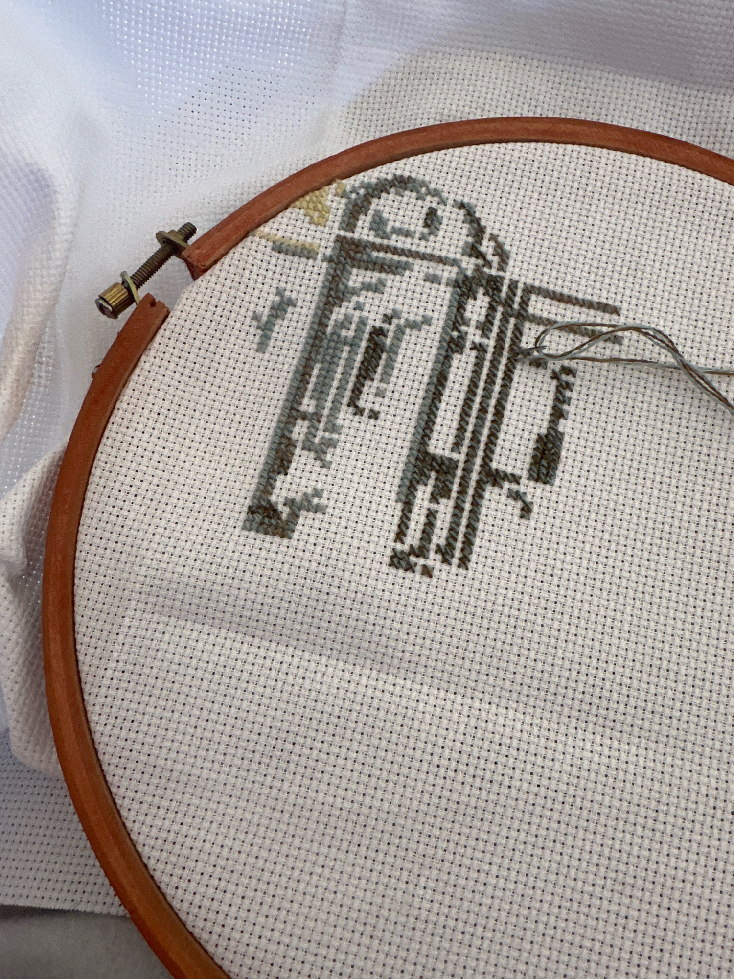The image shows a close-up of a white cross-stitch fabric stretched within a wooden embroidery hoop. The fabric is partially embroidered with a design in shades of grey and dark grey thread, forming geometric shapes that resemble a robot or mechanical figure. A needle threaded with light grey and dark grey thread rests on the fabric, with a portion of the thread extending off the hoop. The embroidery hoop has a metal screw and nut closure, which is slightly visible. The background consists of white fabric, possibly a tablecloth or a larger piece of material, with a grey surface underneath. There are no people or recognizable landmarks of Gyumri, Armenia, visible in the image.