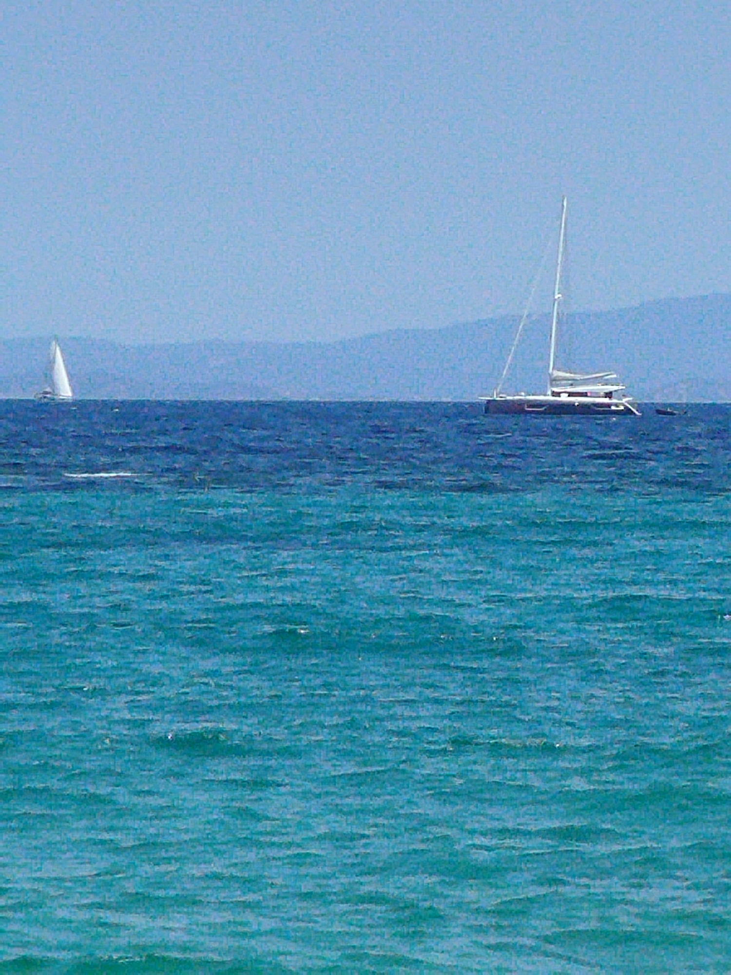 A wide shot captures a body of water with two sailboats in the distance. The water is a vibrant turquoise color and appears choppy with small waves. In the background, a thin, hazy line of land with mountains is visible under a clear, pale blue sky. The sailboat on the left is smaller and closer to the shore, with a single white sail. The sailboat on the right is larger and further out, appearing to be a catamaran with multiple white sails and a dark hull. The image is taken from a slightly elevated perspective, looking out over the water.