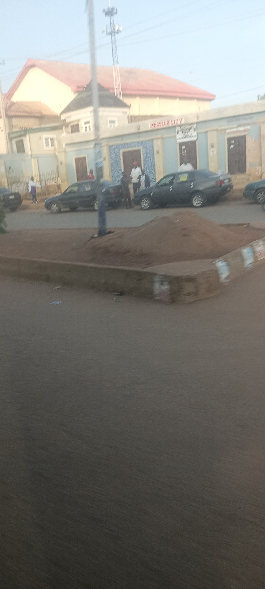 The image depicts an outdoor scene in Bukuru, Nigeria. In the background, a light blue building with multiple doorways and windows is visible, with "MESSIAN CITY" signage above one of the entrances. A larger building with a red roof is situated behind this structure. In the foreground, a street scene unfolds with several dark-colored sedans parked or moving. A mound of dirt is present in the middle ground. Several individuals are visible, some standing near the vehicles and buildings, and one person appears to be walking across the street. The overall lighting suggests it is daytime, possibly late afternoon, given the soft light.