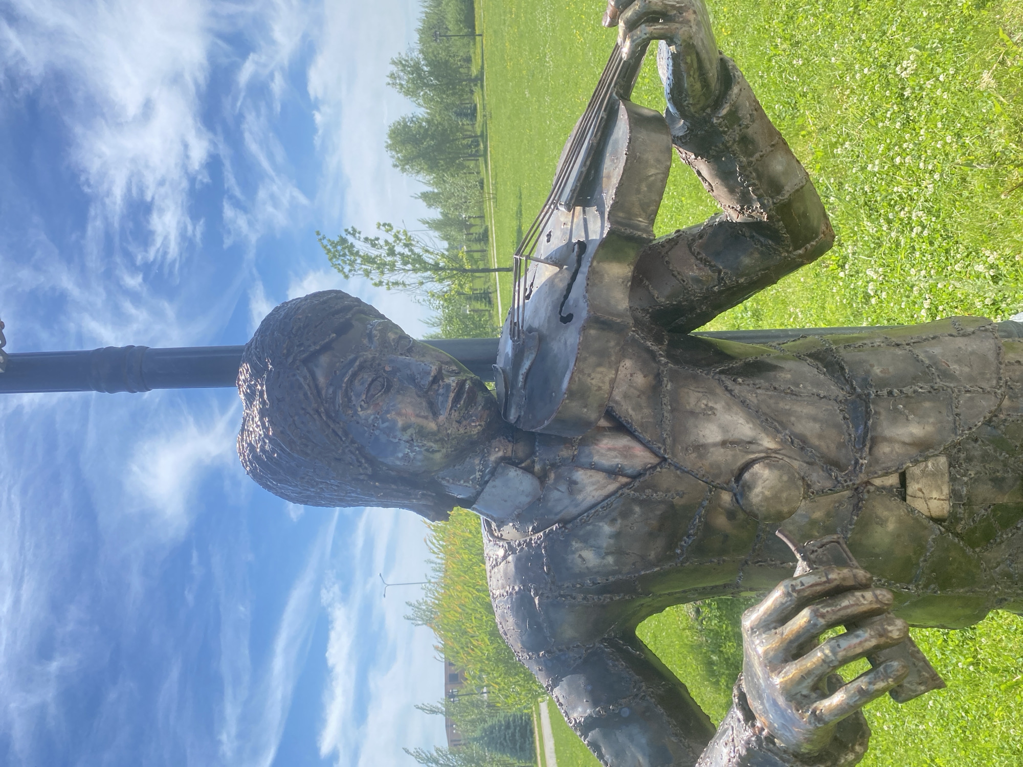 The image captures a unique metallic sculpture of a man playing a violin, situated outdoors in a bright, park-like environment. The sculpture is composed of numerous welded metal plates, giving it a distinctive, segmented appearance and a reflective surface. The man, depicted with a focused expression, holds a metallic violin, complete with visible strings and f-holes, as if engrossed in a performance. His hands are positioned as a violinist's would be, with his left hand on the neck and his right hand holding a bow.
The setting is an expansive green grassy field, interspersed with small white flowers, suggesting a vibrant spring or summer day. A line of deciduous trees frames the middle ground, with additional foliage and hints of distant structures in the background, all bathed in natural sunlight. The sky above is a clear, bright blue, adorned with wispy white cirrus clouds, indicating pleasant weather. A dark, vertical pole is partially visible behind the sculpture's head. The scene evokes a tranquil moment of musical art within an outdoor public space, consistent with a park setting in Tyumen, Russia. There is no visible text in the image.