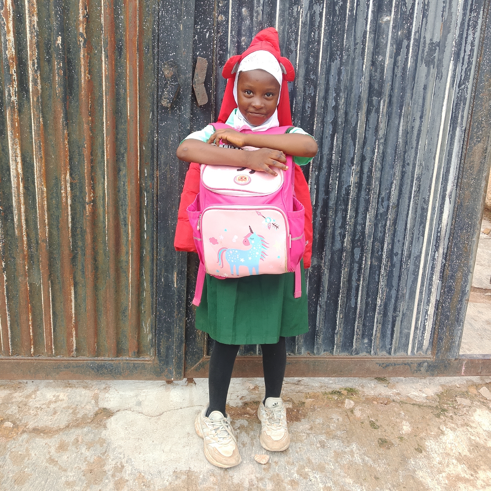 A young girl stands in front of a corrugated metal gate, her arms crossed over a pink backpack. She wears a white hijab and a red costume with ears, and a green school uniform skirt. She has on black tights and light-colored athletic shoes. The gate is dark gray and appears weathered, with patches of rust and dirt. The ground is a light-colored concrete surface with some cracks and debris.