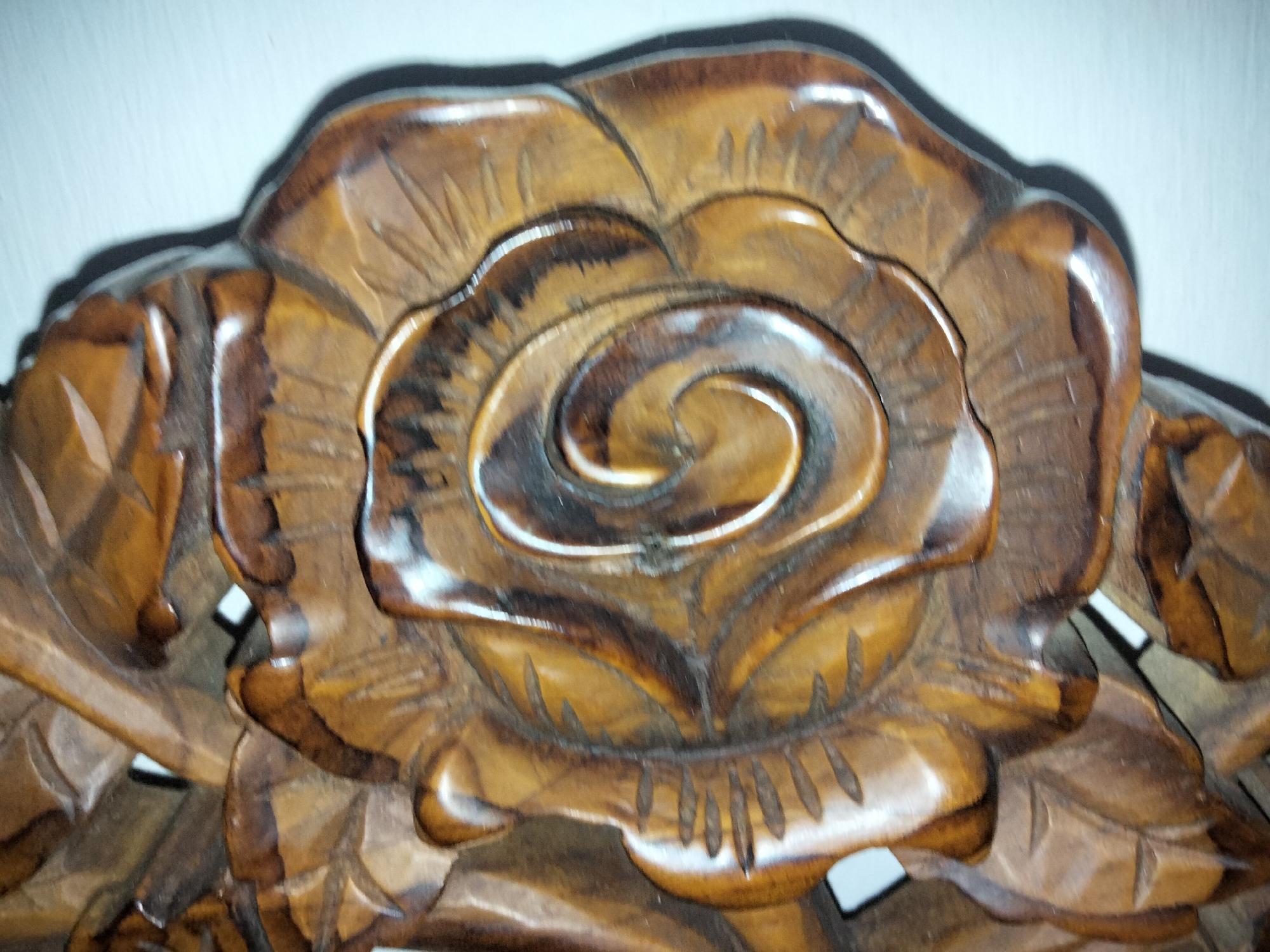 The image shows a close-up, overhead view of a carved wooden rose. The carving is detailed, with visible petal layers and a central swirl. The wood has a glossy finish, reflecting light and highlighting the carving's depth. The rose is part of a larger, decorative carving, with hints of leaves and other floral elements visible around the edges. The background is a plain, light-colored wall, with a shadow cast by the carving, indicating it is mounted or placed against it. The lighting suggests an indoor setting. The image does not contain any information about the city of Payagaboh, Indonesia, or any people.