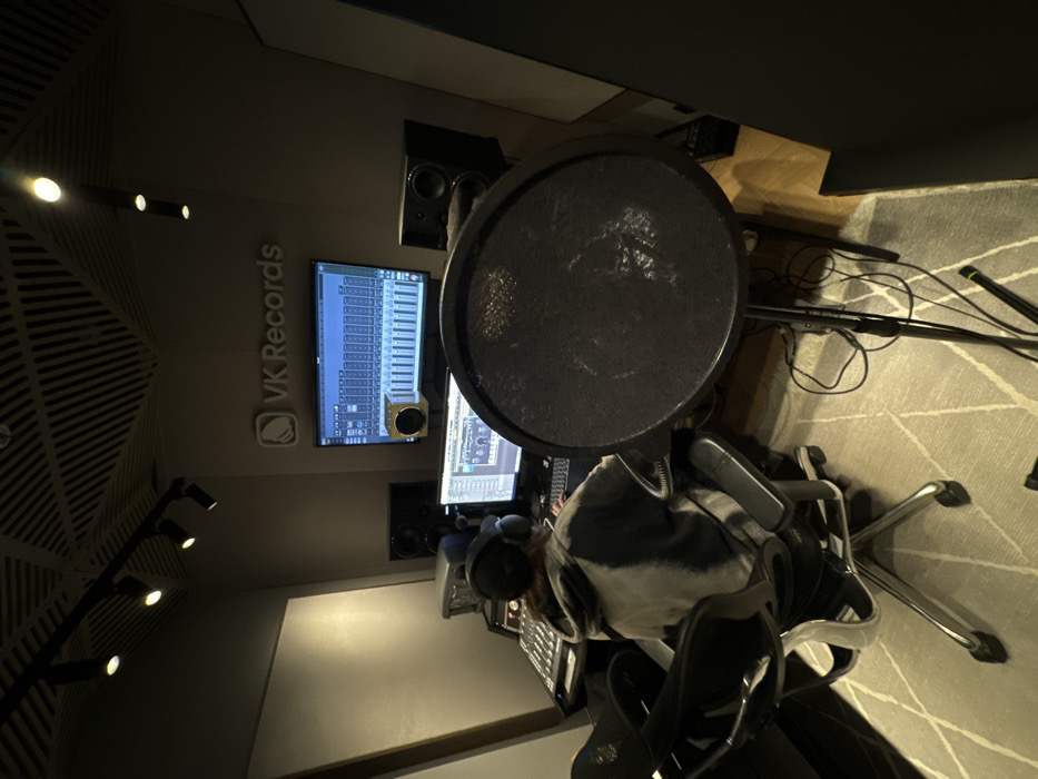 The image depicts the control room of a recording studio, identified by the "VK Records" branding displayed on the wall above a primary computer monitor. This studio is located at ulitsa Mayakovskogo, 2Б, Khimki, Moscow Oblast, Russia. A single individual is depicted from the rear, seated in a black ergonomic office chair with chrome accents. The person wears a grey and white tie-dye hooded sweatshirt and dark over-ear headphones, and is operating a keyboard. Two computer monitors are visible; the larger upper screen displays digital audio workstation (DAW) software with multiple tracks and a virtual keyboard, while a smaller monitor below shows additional software. To the left of the individual, an audio mixing console is present. Studio monitor speakers are positioned on either side of the main display, with the left speaker resting on a yellow isolation pad. In the foreground, a black circular pop filter is mounted on a stand, with associated cabling on the floor. The studio environment features grey acoustic panels on the walls and a slatted wooden acoustic ceiling. The floor is covered by a light-toned geometric patterned rug, revealing some wooden flooring. Track lighting fixtures illuminate the space.