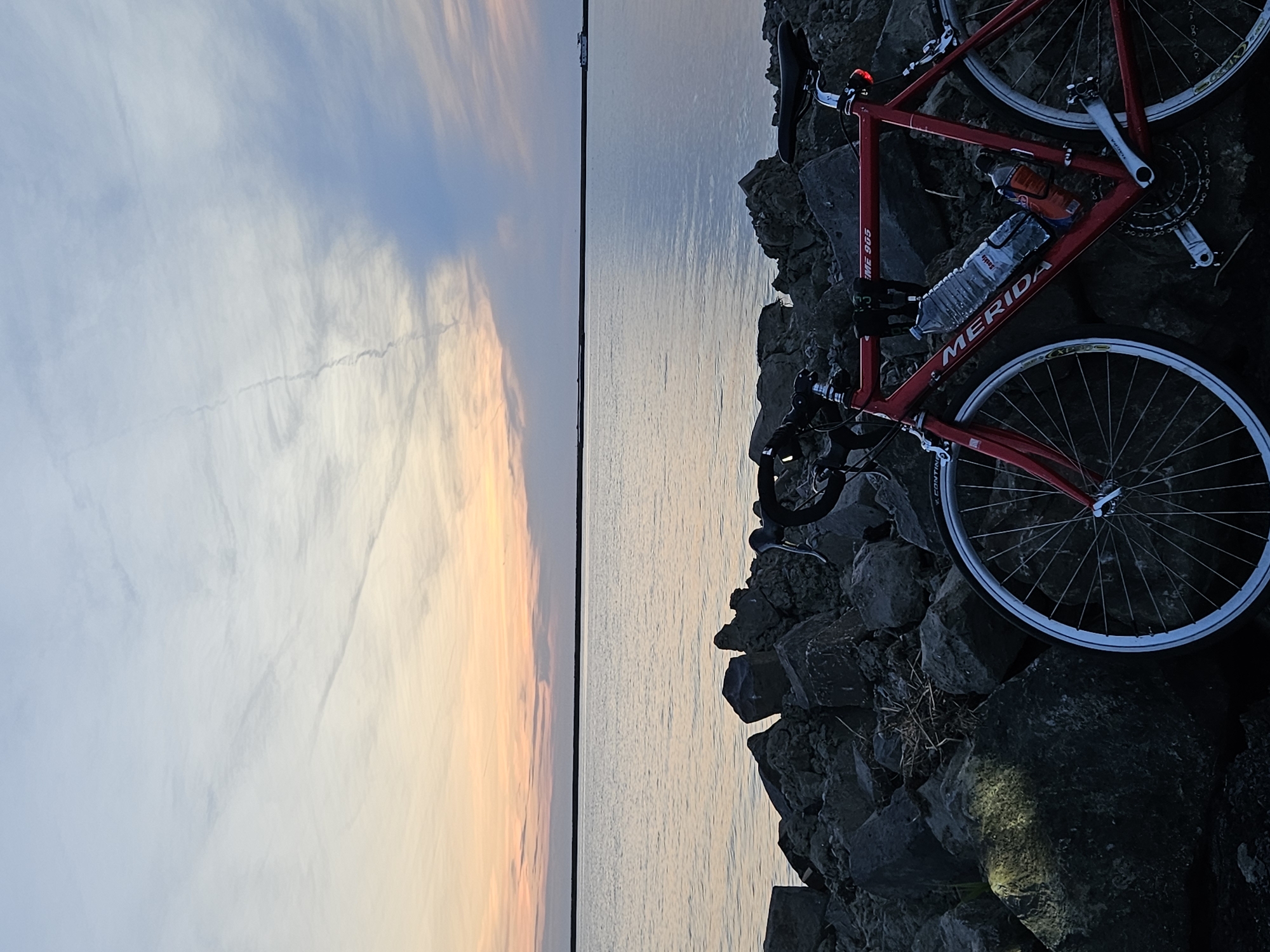 A red Merida bicycle is parked on a rocky embankment beside a body of water. The bicycle is facing towards the right of the frame and has two water bottles attached to the frame. The water appears calm, reflecting the soft colors of a sunset sky. The sky transitions from pale orange and pink near the horizon to a pale blue and white higher up, with wispy clouds visible. In the distance, on the water, a small white structure, possibly a boat or a building, is visible. The rocky embankment is composed of dark grey stones of various sizes. The scene is set at dusk. The location is not explicitly identifiable from the image alone, but if this image is indeed from Lelystad, Netherlands, it could be along the IJsselmeer or one of its surrounding waterways.