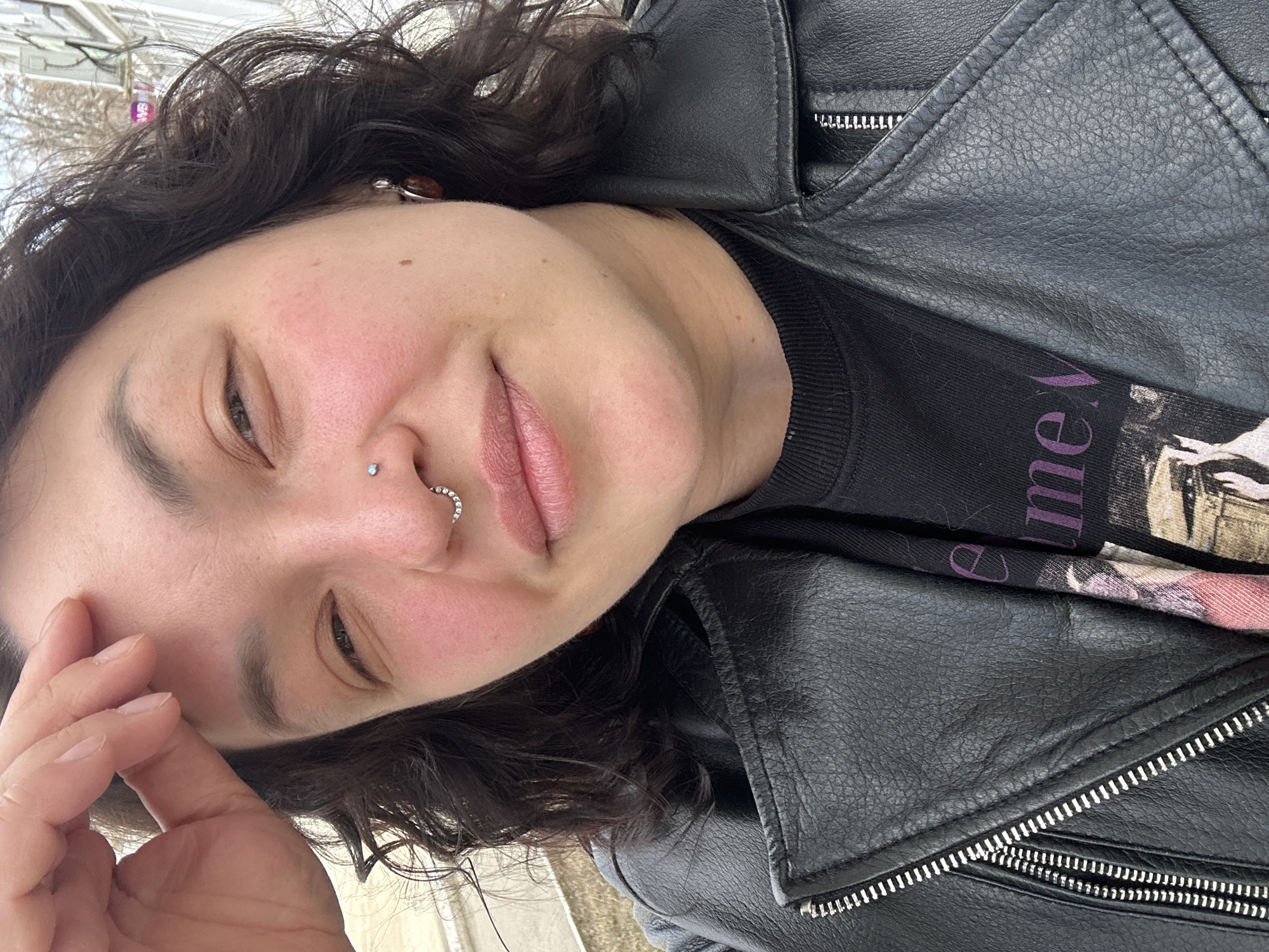 A close-up, tilted portrait captures a person with dark, wavy hair, likely outdoors in Chaikovsky, Russia. The individual, appearing to be a young adult, looks towards the viewer with a relaxed, slightly smirking expression. Their cheeks have a natural, rosy flush, and their lips are a soft pink. Notable facial features include two nose piercings: a small, iridescent stud on the right nostril and a ring adorned with tiny, shimmering stones on the left. One hand is visible in the lower left foreground, with fingers gently resting near their forehead.
The person is dressed in a black leather jacket featuring prominent silver zippers, worn over a dark-colored, possibly black, t-shirt. On the t-shirt, a fragment of text in purple is visible, appearing as "ne W" or "me V," along with some faint, lighter-toned graphical elements. The background is softly blurred, indicating an outdoor setting during daytime, with indistinct light-colored structures and hints of an open sky or light foliage creating a diffuse backdrop. The overall impression is casual and candid.