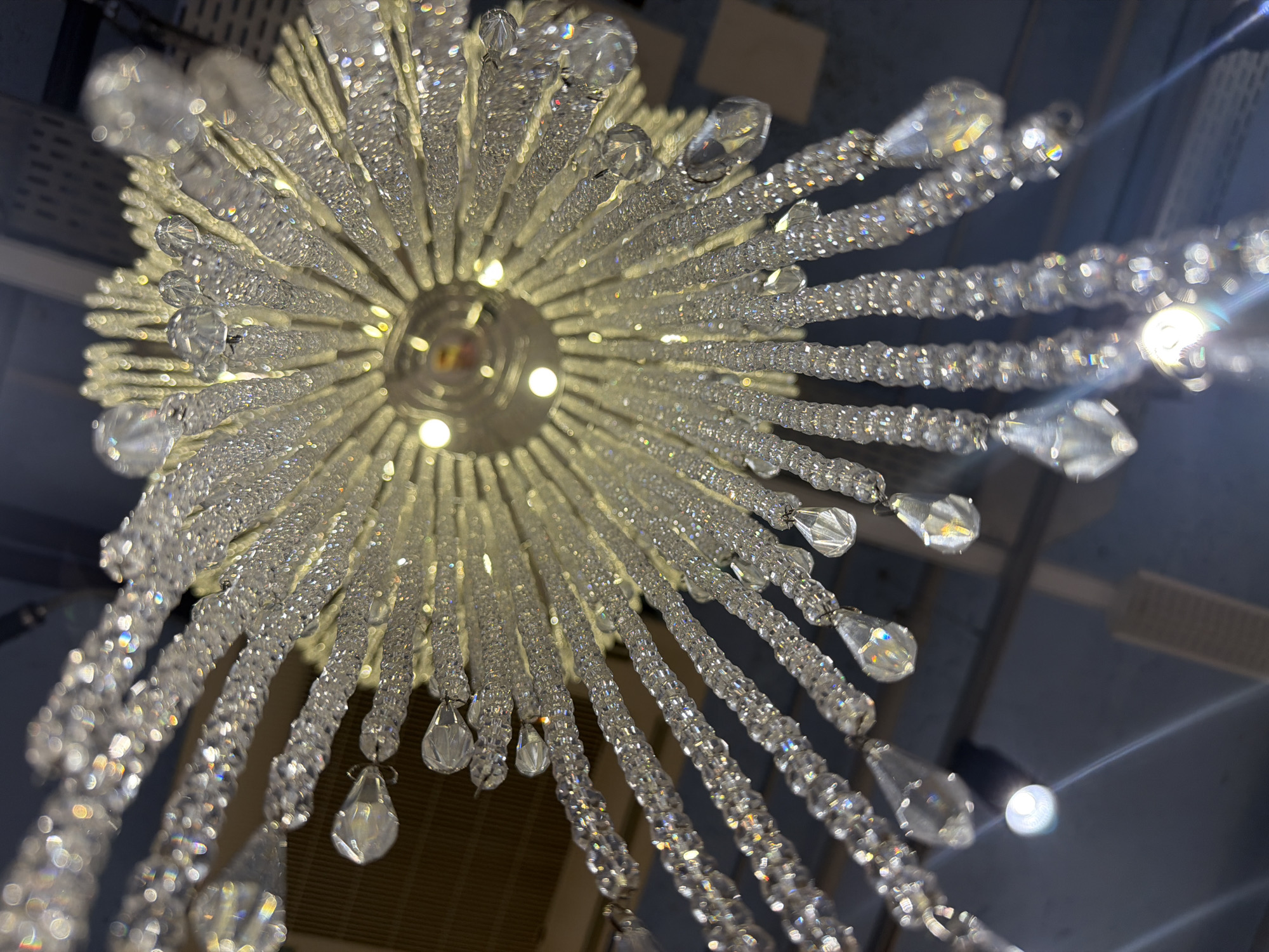 A low-angle shot depicts a crystal chandelier. The chandelier consists of many strands of small, clear beads radiating outward from a central hub. The beads reflect light, creating a shimmering effect. Some strands end with larger, teardrop-shaped crystals. The chandelier is likely in a commercial or residential building. The image was taken in the city of Spreitenbach, Switzerland.