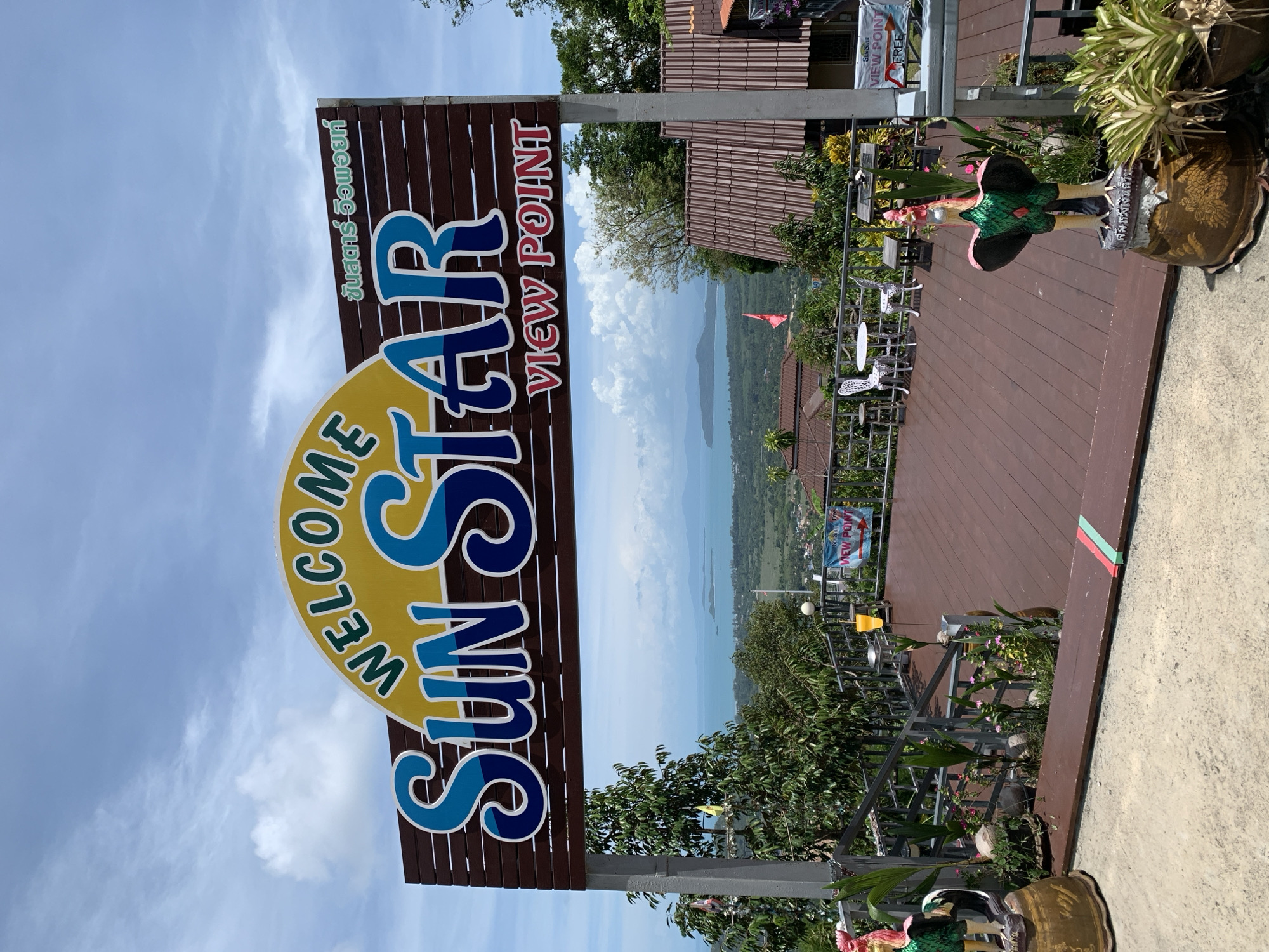 The image shows a sign that reads "WELCOME SUN STAR VIEW POINT" at the Sun Star View Point in Baan Thurian, Thailand. The sign is a large, wooden structure with the text printed in a bold, colorful font. The view point overlooks a lush, green landscape and the ocean. There is a deck with outdoor seating and foliage surrounding the area. The sky is partly cloudy, with some blue visible. The image is rotated to the right, with the sign and the view point partially obscured.