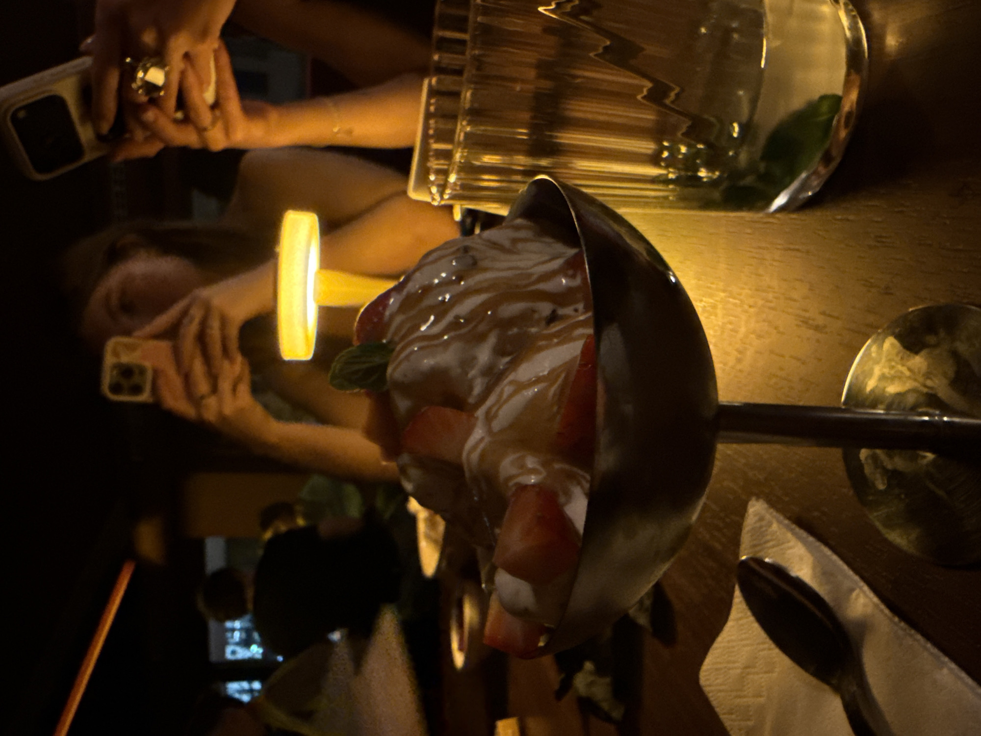 A dessert, consisting of strawberries and cream in a black bowl on a silver stand, is on a brown table with a glass of water and a spoon to the right. Reflected in the table are two people holding phones, with one person’s hand visible in the upper left corner with a large ring. A straw is visible on the lower left side. The scene is likely in Belgrade, Serbia.