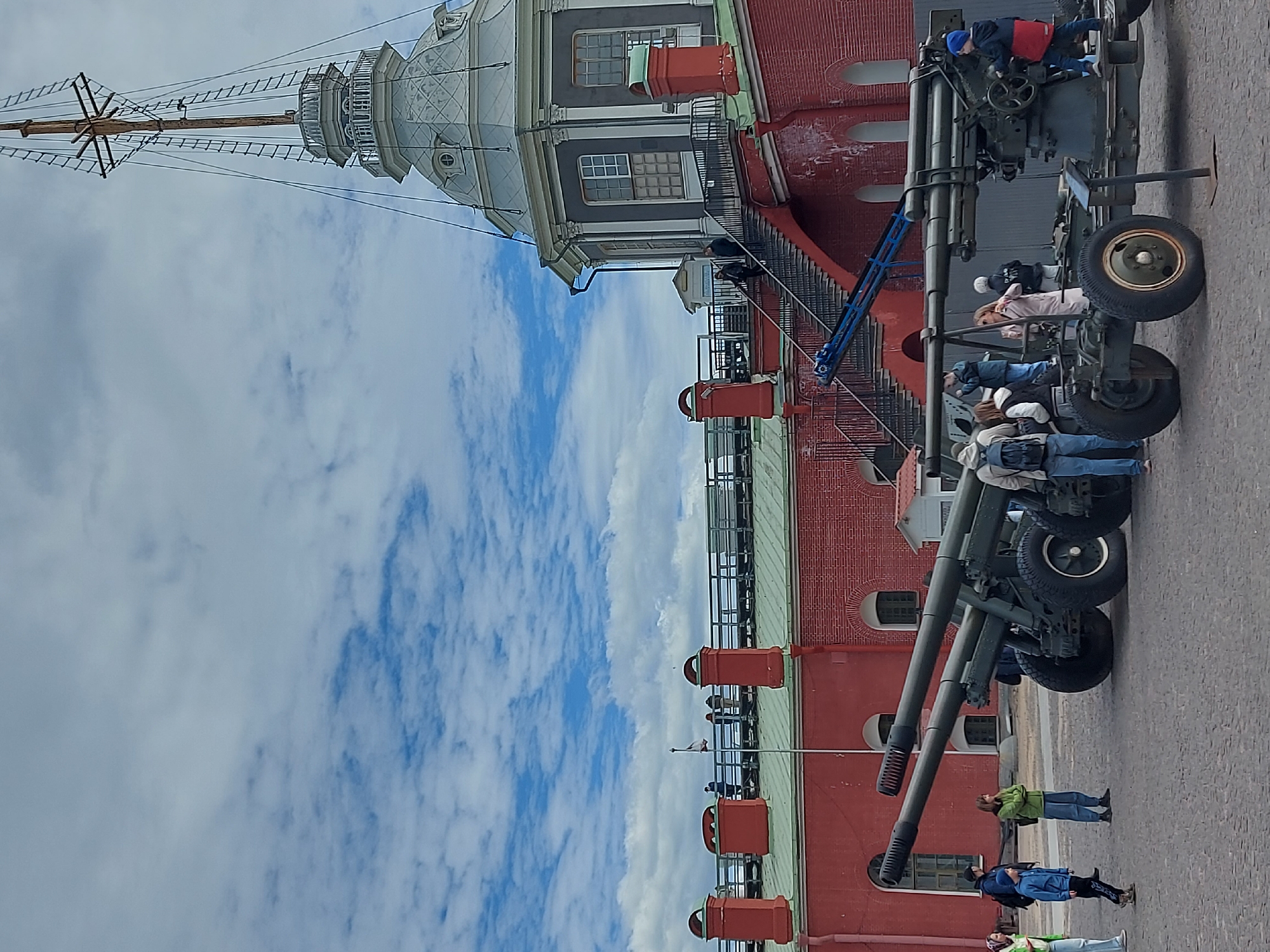 Here is a detailed description of the image:
The image presents a scene at the Peter and Paul Fortress in Saint Petersburg, Russia. The frame features the iconic fortress cathedral spire against a partly cloudy sky, and its red brick walls.
On the right of the frame are several large artillery guns on wheeled carriages. People are shown interacting with the artillery. Some are sitting or climbing on the guns, while others are standing nearby, presumably observing or taking photos.
The environment appears to be a paved courtyard or open space within the fortress. The overall impression is that of a historical site visited by tourists.