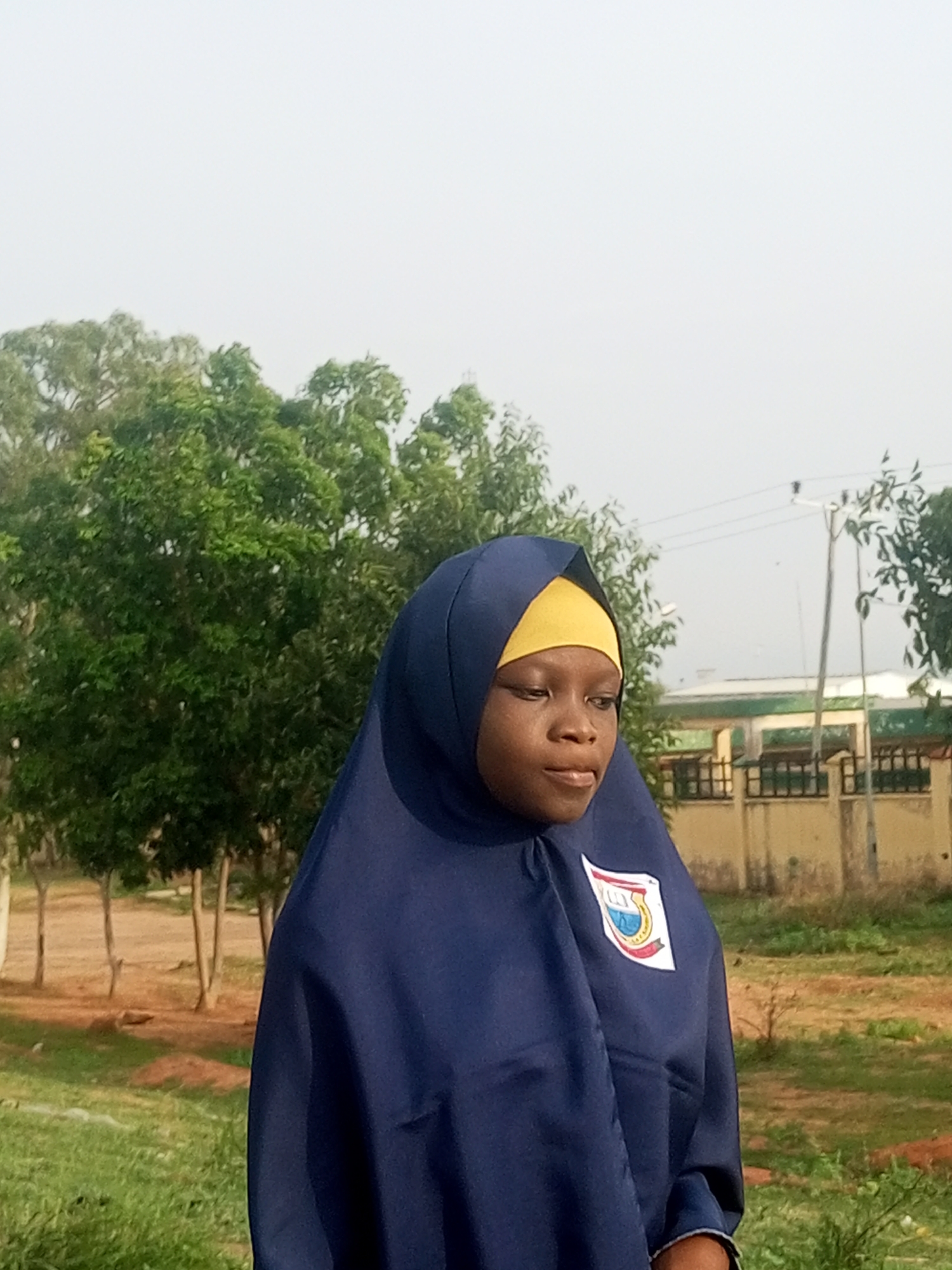 A medium shot captures a young girl in a navy blue hijab and yellow headscarf, looking to her right with a slight smile. She wears a navy blue garment with a patch on the left side of her chest, featuring a white, red, and blue emblem with text. Her left hand is visible, partially obscured by her hijab. She is outdoors, with a background of lush green trees, a dirt path, and a building with a tan wall and green trim. The sky is a hazy, pale blue. The lighting suggests it is daytime.
