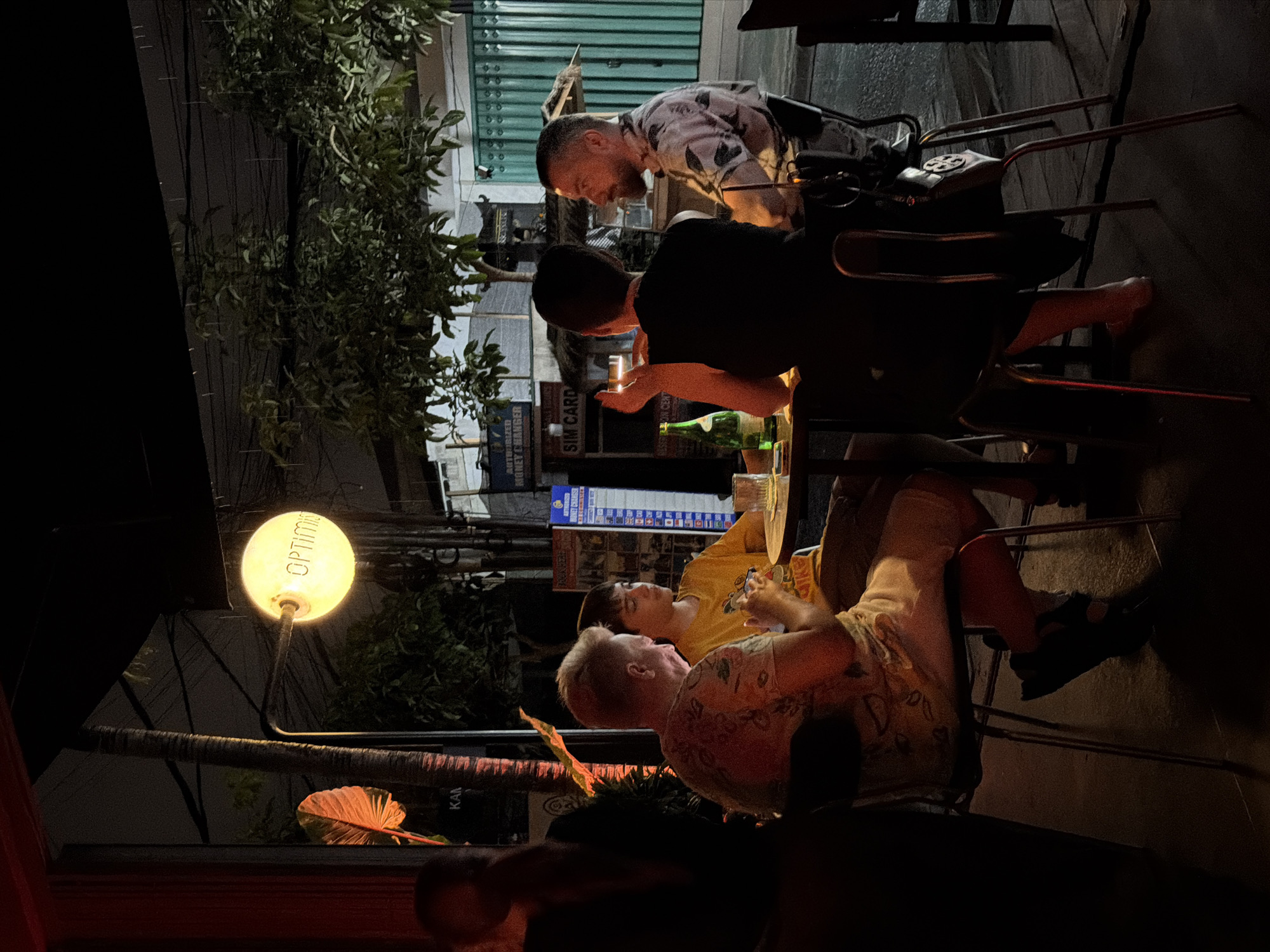 The image captures a nighttime scene at what appears to be an outdoor seating area, possibly a restaurant or bar, in Mengwi, Indonesia. Several individuals are present, seated at tables and chairs. The lighting is dim, with a warm, yellow glow emanating from a spherical lamp that reads "OPTIM..." visible on the left side of the frame.
Two men are prominently featured in the foreground, seemingly engaged in conversation or an activity at a table. One man, wearing a patterned shirt, leans back in his chair, while another man, in a black t-shirt, leans forward. Further back, another man in a white and pink patterned shirt is visible. A fourth person, wearing a yellow t-shirt, is seated and looking towards the foreground.
The setting is characterized by lush greenery from an overhanging plant on the left, contrasting with the man-made structures of tables, chairs, and what appears to be a building or facade in the background. Some signage is partially visible, including a "SIM CARD" sign and another with what looks like a price list or menu. The overall atmosphere is relaxed and casual, suggesting a social gathering or a moment of leisure. The presence of overhead lighting and the general ambiance indicate it is nighttime.