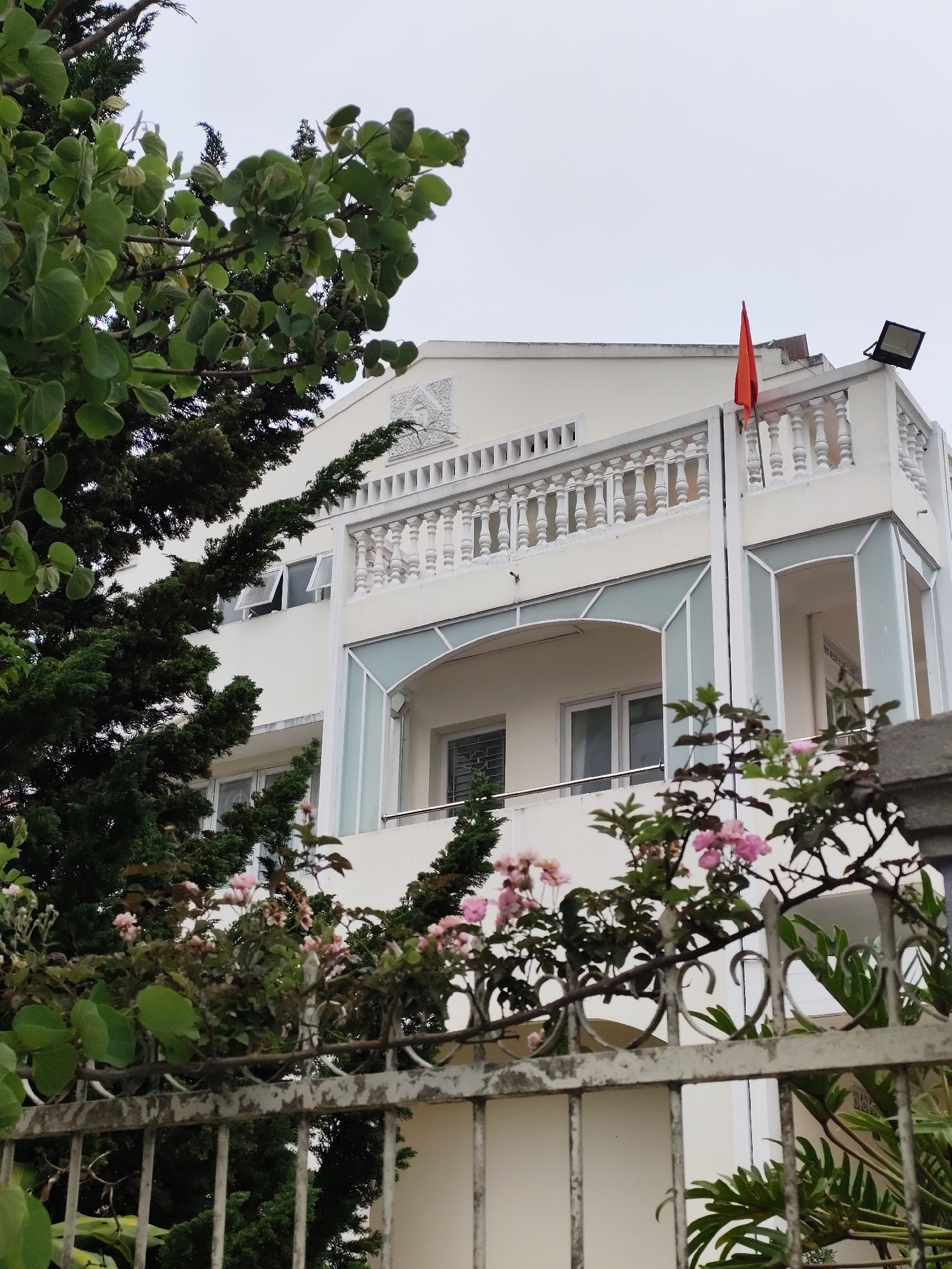 The exterior of a white, multi-story building is partially obscured by greenery and a metal fence. The building has a prominent balcony with a balustrade and an orange flag. A light fixture is visible on the roof. The scene is in Da Lat, Vietnam. The weather is overcast. No people are visible.