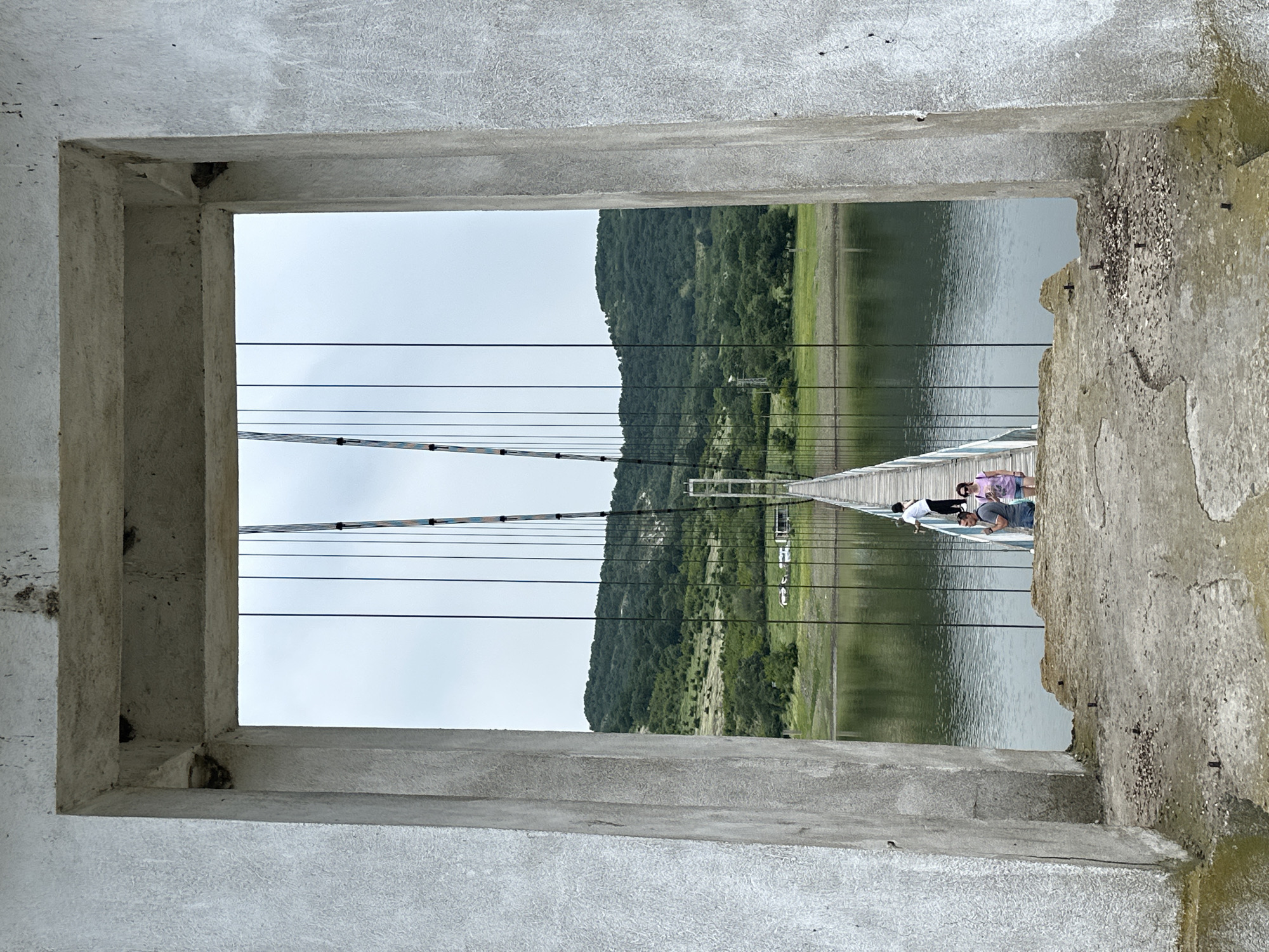 The image depicts a concrete rectangular archway framing a view of the Lisitsite suspension bridge spanning a body of water, likely a reservoir, in Lisitsite, Bulgaria. The archway, made of rough, light grey concrete, shows signs of weathering and minor spalling, with exposed rebar stubs embedded in the concrete ground in the immediate foreground.
Through the archway, the narrow suspension bridge extends across calm, dark green-grey water. The bridge features a wooden plank walkway, white metal railings, and numerous dark vertical suspension cables connected to larger overhead cables. Two main bridge towers are visible, with the closer tower's cables displaying colorful markings.
Three individuals are present on the bridge, walking away from the viewer towards the far bank. The furthest individual is a person in a white shirt and dark pants, positioned on the right side of the bridge. Closer to the viewer, a man in a grey t-shirt and blue shorts walks alongside a woman in a pink t-shirt and patterned shorts, both on the left side of the bridge.
The background consists of rolling hills covered densely with green deciduous trees and lighter green scrubland. On the far bank, near the bridge's end, a few vehicles, including a white car and a camper, are discernable. The sky above is overcast and light grey.