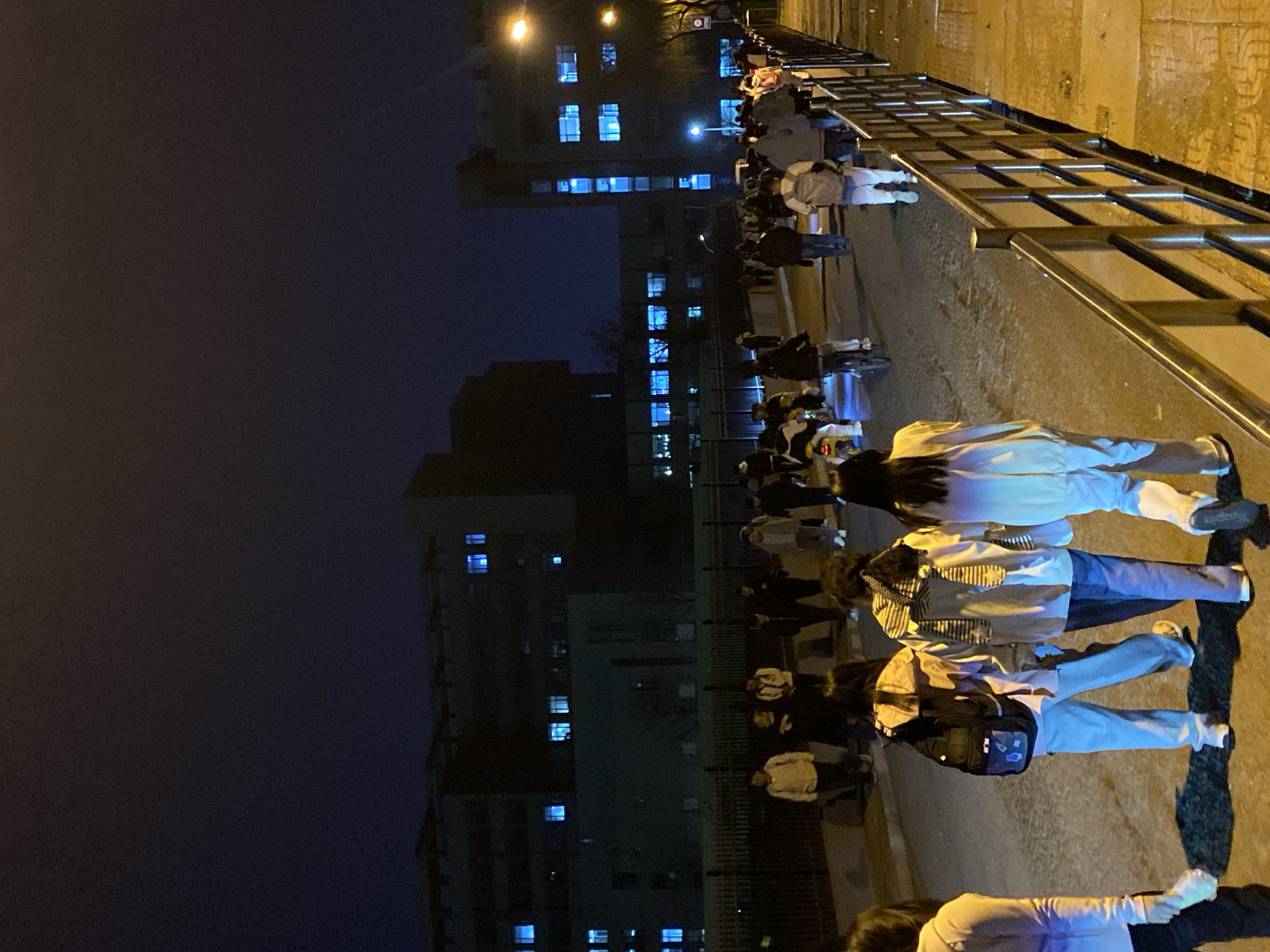 A nighttime view in what appears to be Zhongtun, China, depicts a group of people walking along a sidewalk next to a metal railing. A tall building with illuminated windows is visible across the street. The sky is dark, and the street appears to be lit by overhead lighting. The people are dressed in casual clothing.