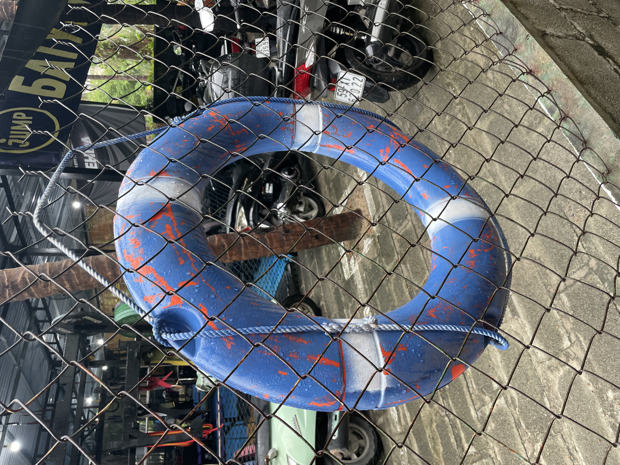 A blue and white life preserver hangs on a chain-link fence in Phu Thuy Ward, Vietnam. Behind the fence, motorcycles are parked. A sign reading "JUMP" and part of the word "BALI" is visible to the left of the preserver. The ground is concrete.