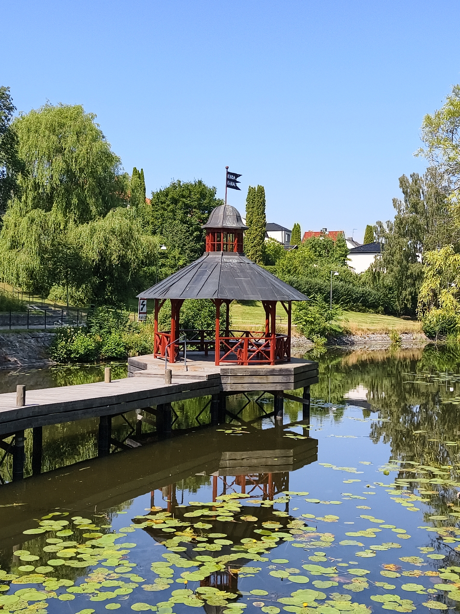 The image depicts a picturesque scene at Kindamossen pond, a scenic spot located in Linköping, Sweden. A wooden boardwalk extends from the foreground into the water, leading to a charming gazebo situated on a small floating platform. The gazebo features a dark, octagonal roof supported by red wooden pillars, with an open-air design allowing views of the surrounding greenery. A small dark flag with white text, possibly indicating a location or name, is visible atop the gazebo's spire. The pond itself is calm and reflects the gazebo, boardwalk, and the lush trees that line the banks. Lily pads are scattered across the water's surface, adding a touch of natural beauty. In the background, a gentle slope is covered with manicured grass, and houses with red and white roofs peek through the dense foliage, suggesting a suburban or park-like setting within Linköping. The sky is a clear, vibrant blue, indicating a sunny day. No people are present in the image.