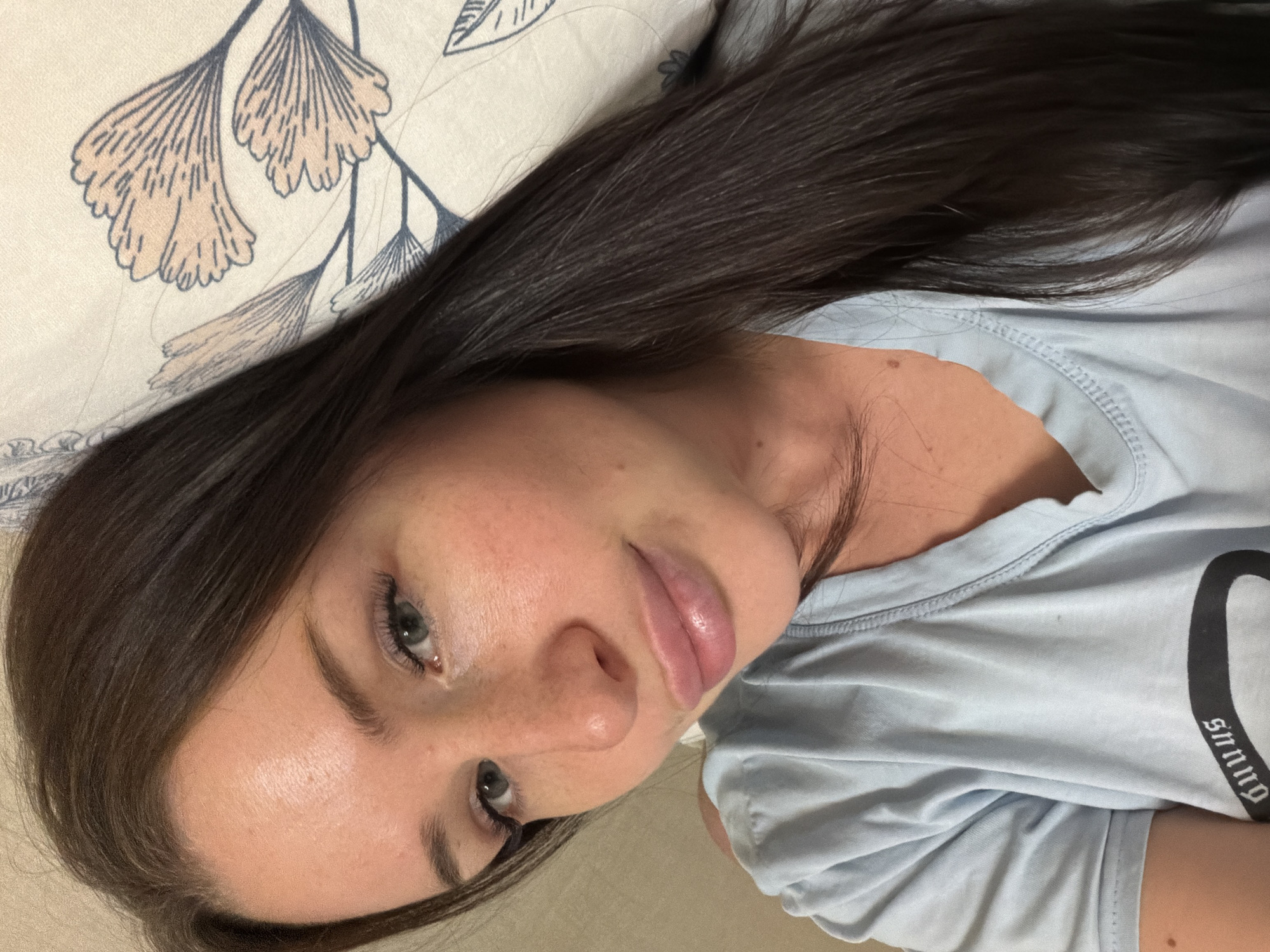 The image displays a young woman with long, straight, dark brown hair, lying down and looking towards the camera. Her head rests on a light-colored pillow or bedding adorned with a repeating pattern of stylized ginkgo-like leaves in light pink/brown and dark blue/grey outlines. She is wearing a light blue t-shirt.
The woman has fair skin with visible freckles across her nose and cheeks. Her eyes are light-colored, appearing blue or grey, and she has neatly shaped eyebrows. Her lips are full. Her expression is calm and relaxed, suggesting a state of rest or quiet contemplation.
A portion of dark grey/black text in a stylized, gothic-style font is visible on the side of her light blue t-shirt, appearing to spell "SnnnQ" or a similar sequence when read from its rotated orientation. The indoor setting, likely a bedroom given the bedding, implies a personal and comfortable atmosphere. The photo is taken from a slightly tilted perspective. This image was captured in Saint Petersburg, Russia.